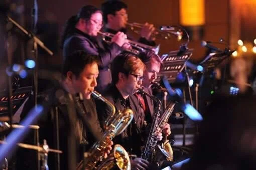A jazz band practicing with five members playing various instruments including saxophones and wind instruments in a dimly lit room.