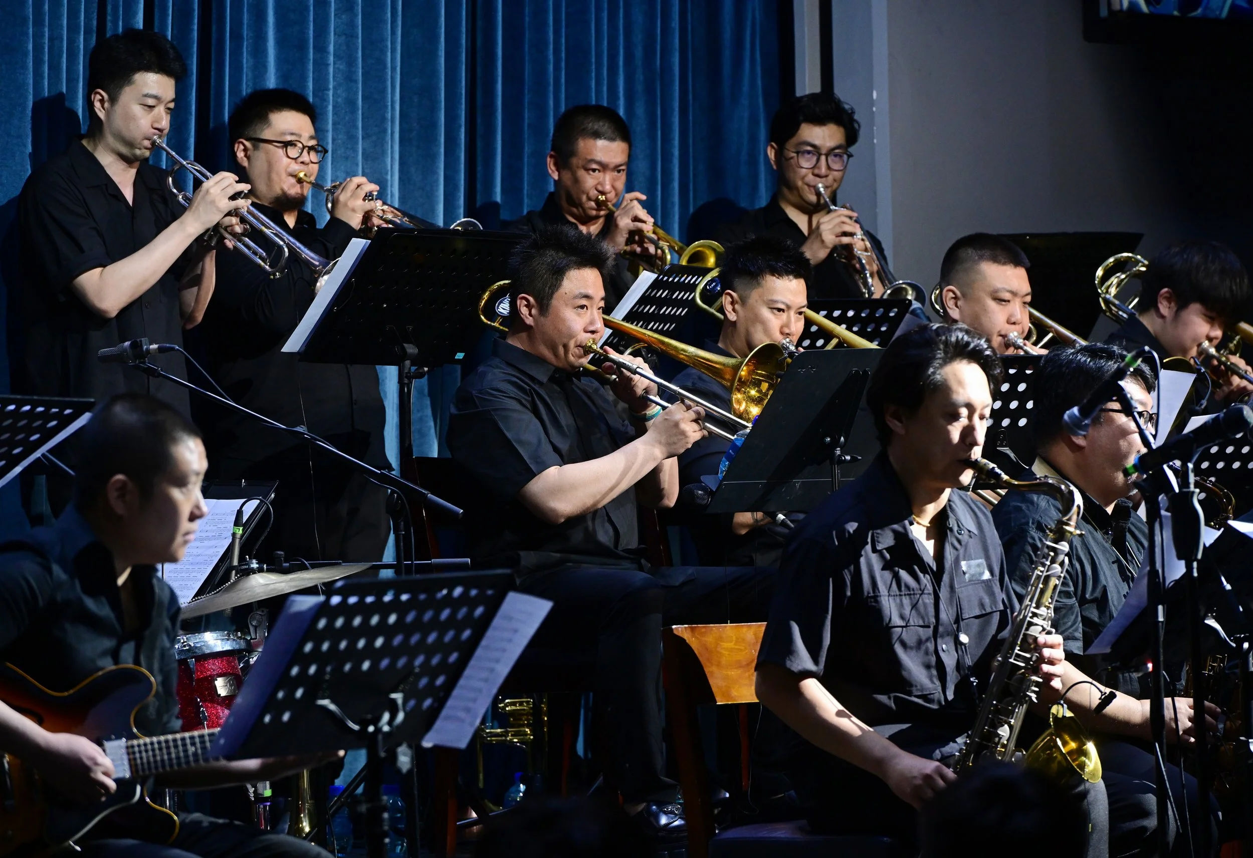 Musicians playing wind instruments and guitar during a performance on stage with blue curtains in the background.
