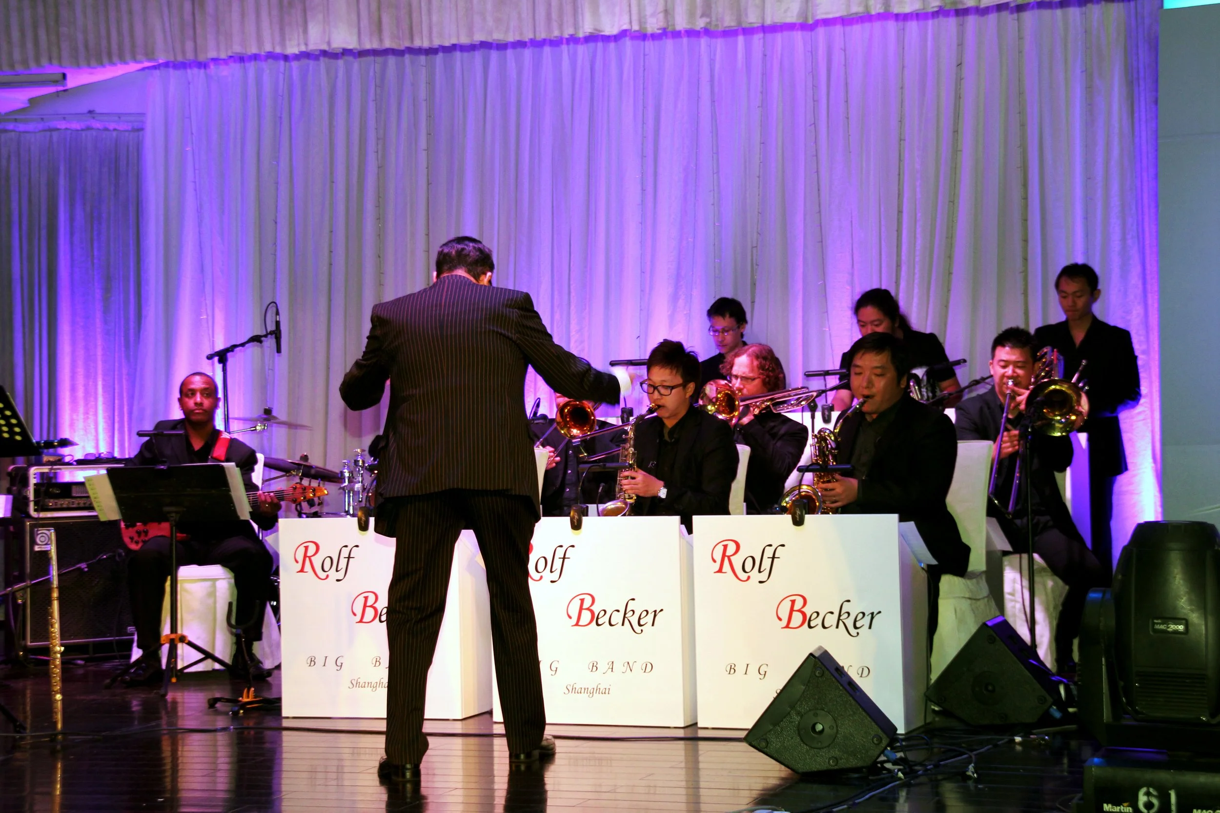 An orchestra performing on stage with a conductor, multiple musicians playing brass instruments, and a backdrop of curtains illuminated with purple lighting. The music stands display the words 'Rolf Becker' and 'BIG BAND Shanghai'.