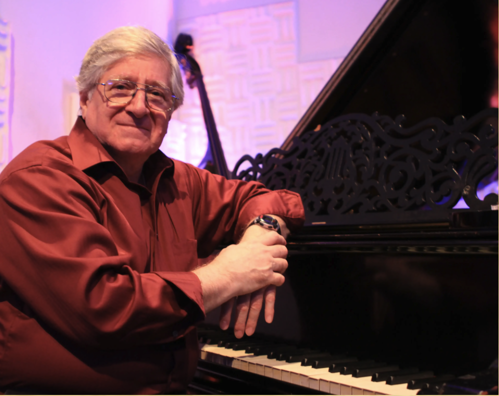 An elderly man with gray hair and glasses wearing a red shirt, sitting at a grand piano with a decorative music stand, in a music studio or performance space.