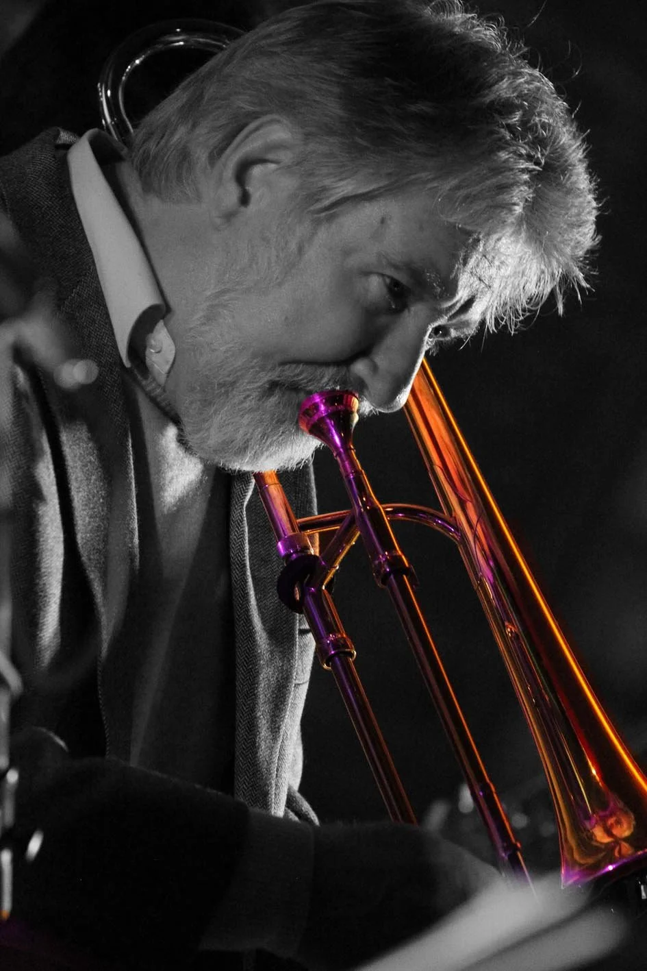 A black and white photo of an older man with gray hair and a beard playing a brass instrument, possibly a trombone, which is illuminated with vibrant purple and orange lights.