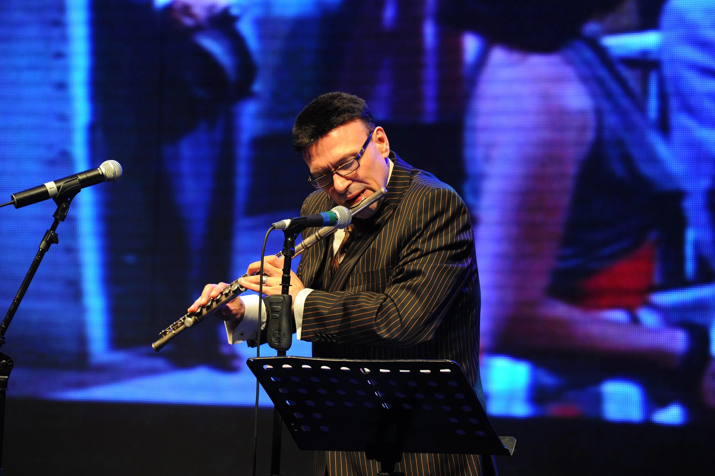 A person with short dark hair and glasses is playing a flute on stage, with microphones in front of them and a large screen displaying abstract visuals in the background.