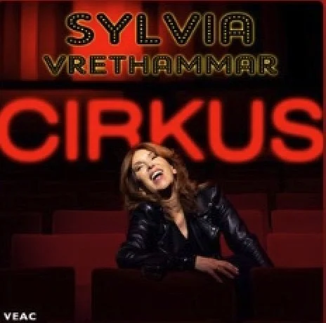 A woman sitting in a theater with a bright red neon sign that reads 'CIRKUS' in the background, and a marquee above with her name 'Sylvia Vrethammar'.