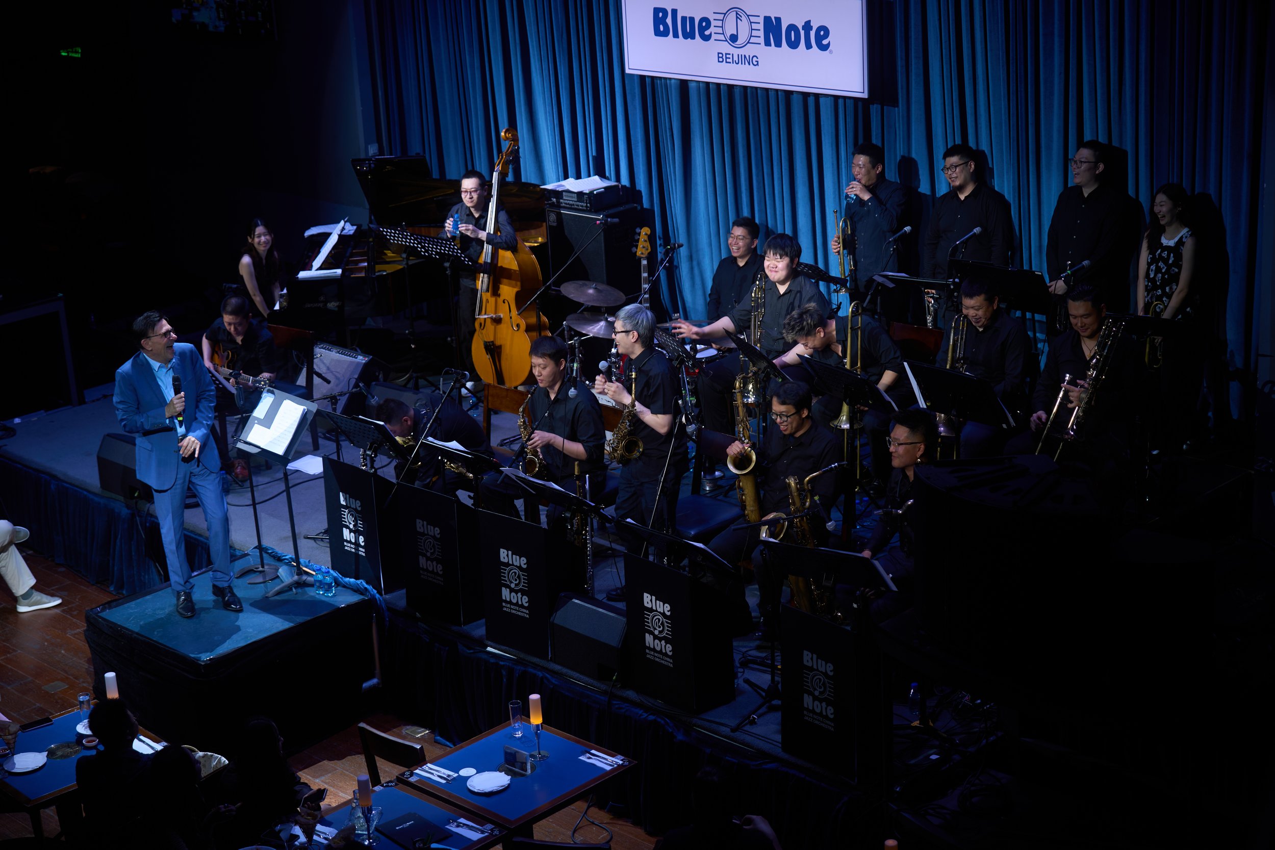 A jazz band performing on stage at Blue Note Beijing, with a singer in a blue suit holding a microphone, various musicians playing instruments including drums, bass, saxophones, and a pianist. The stage has blue curtains and a large sign with the Blue Note logo and the word 'BEIJING'. Audience members are seated at tables with drinks and candles.
