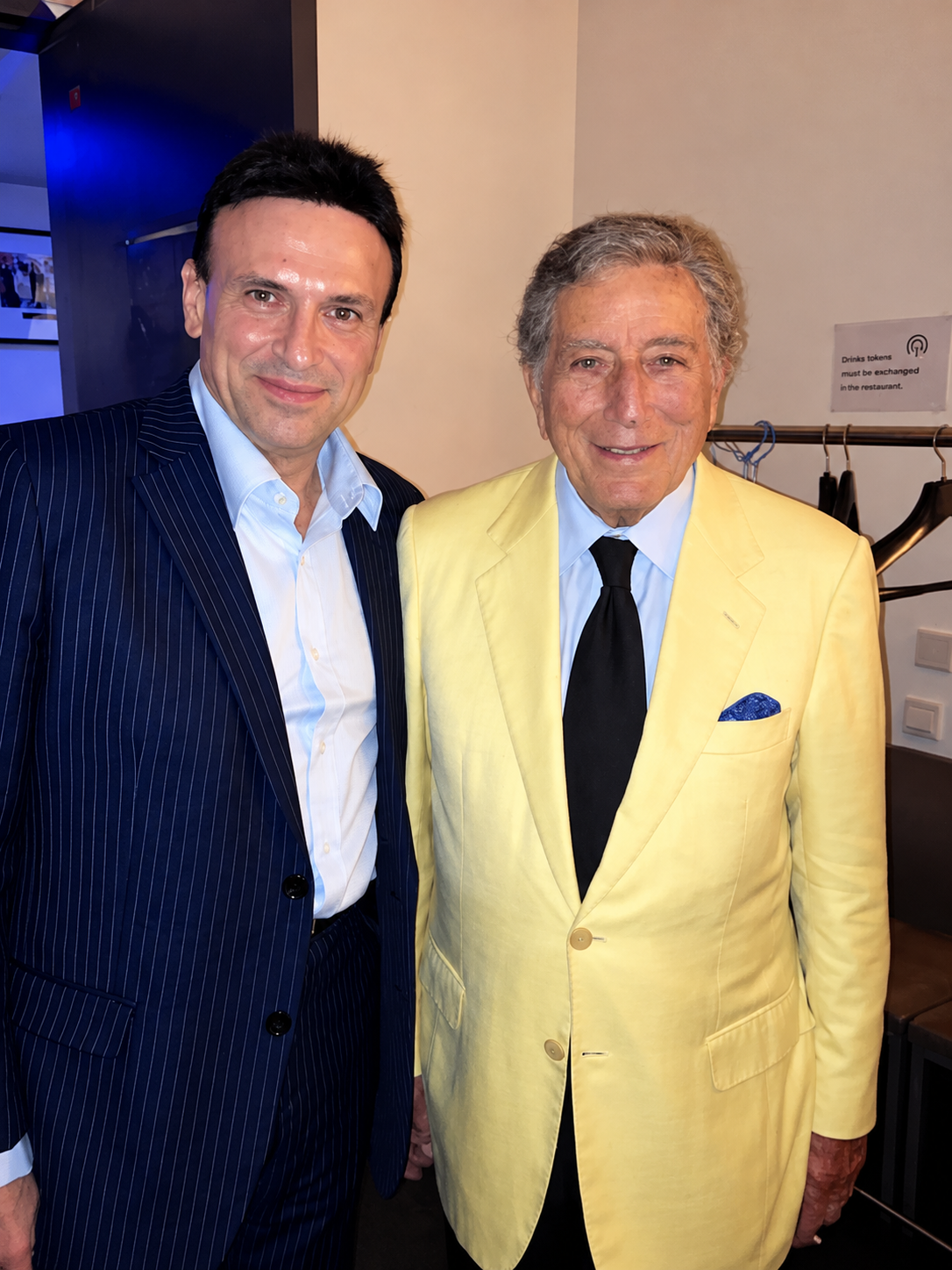 Two men in suits standing together, smiling at the camera. The man on the left wears a dark pinstripe suit with a light blue shirt. The man on the right wears a yellow suit with a light blue shirt, black tie, and a blue pocket square. There is a coat rack with black jackets behind them and a wall with a sign about exchanging drinks tokens.