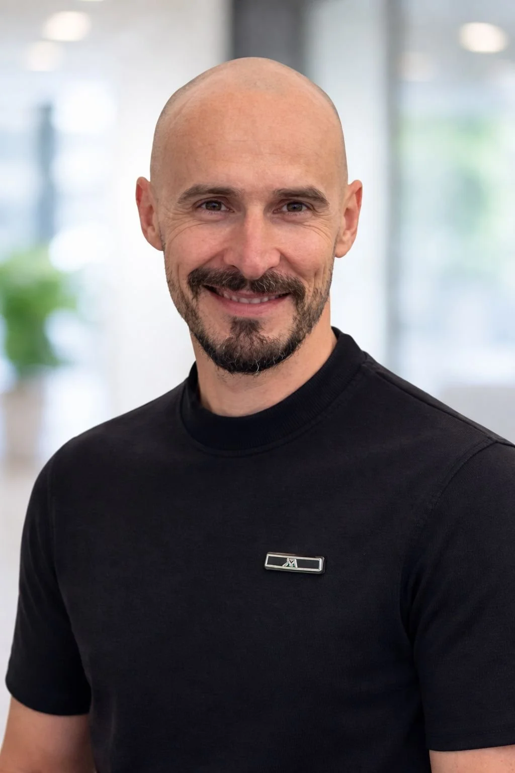 A smiling man with a bald head, beard, and mustache wearing a black t-shirt with a small badge on the chest, standing indoors with a blurred background.