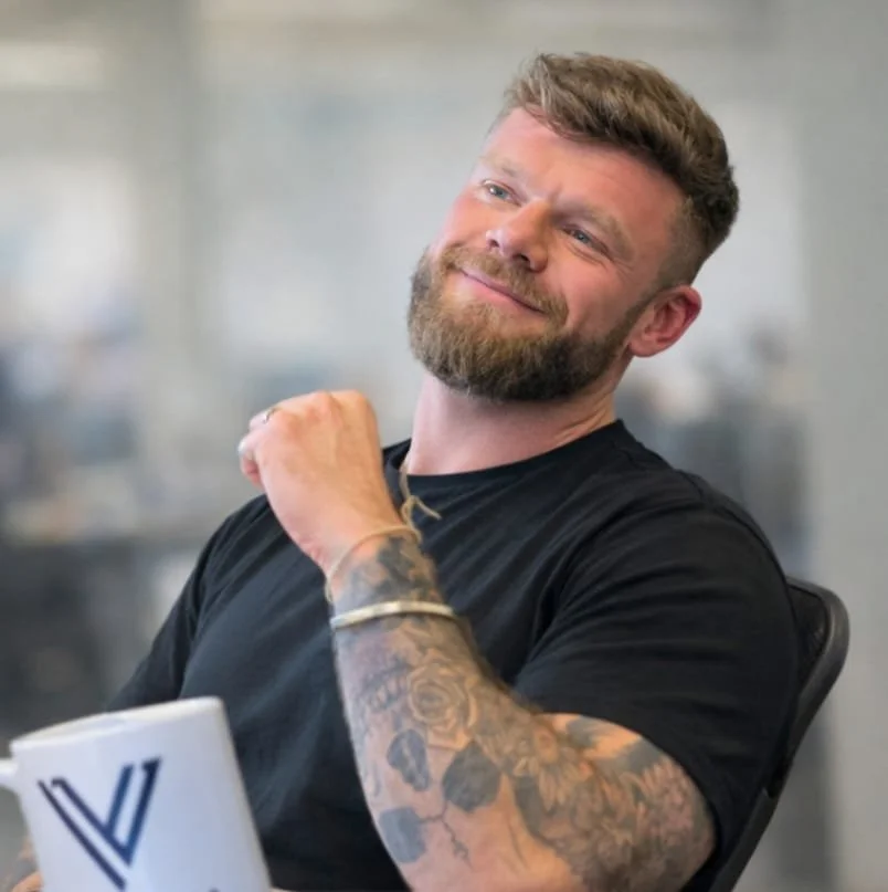A man with a beard and tattoos on his arm is smiling and sitting at a desk in an office, holding a coffee mug in front of him.