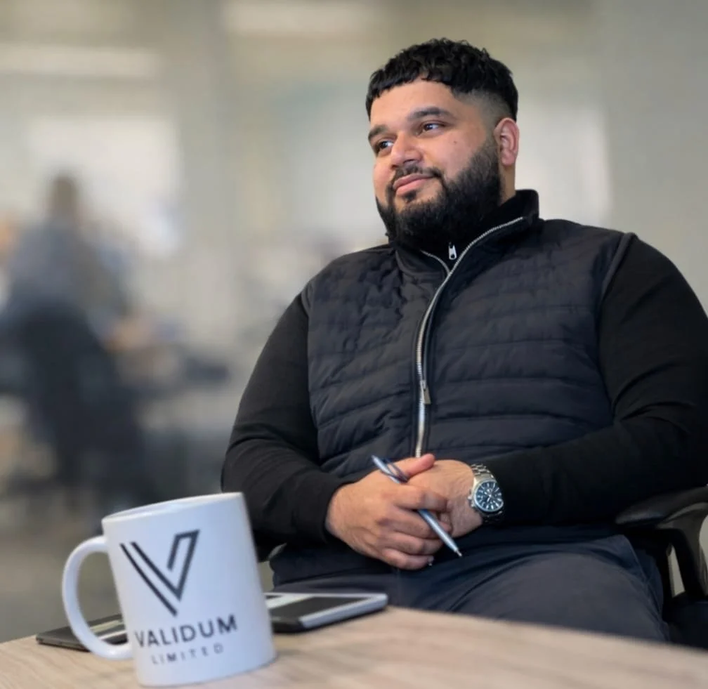 A man with dark hair and a beard, wearing a black quilted vest over a long-sleeve shirt, sitting at a desk with a cup that has the logo 'Validum Limited'. He is holding a pen and appears to be in an office environment.