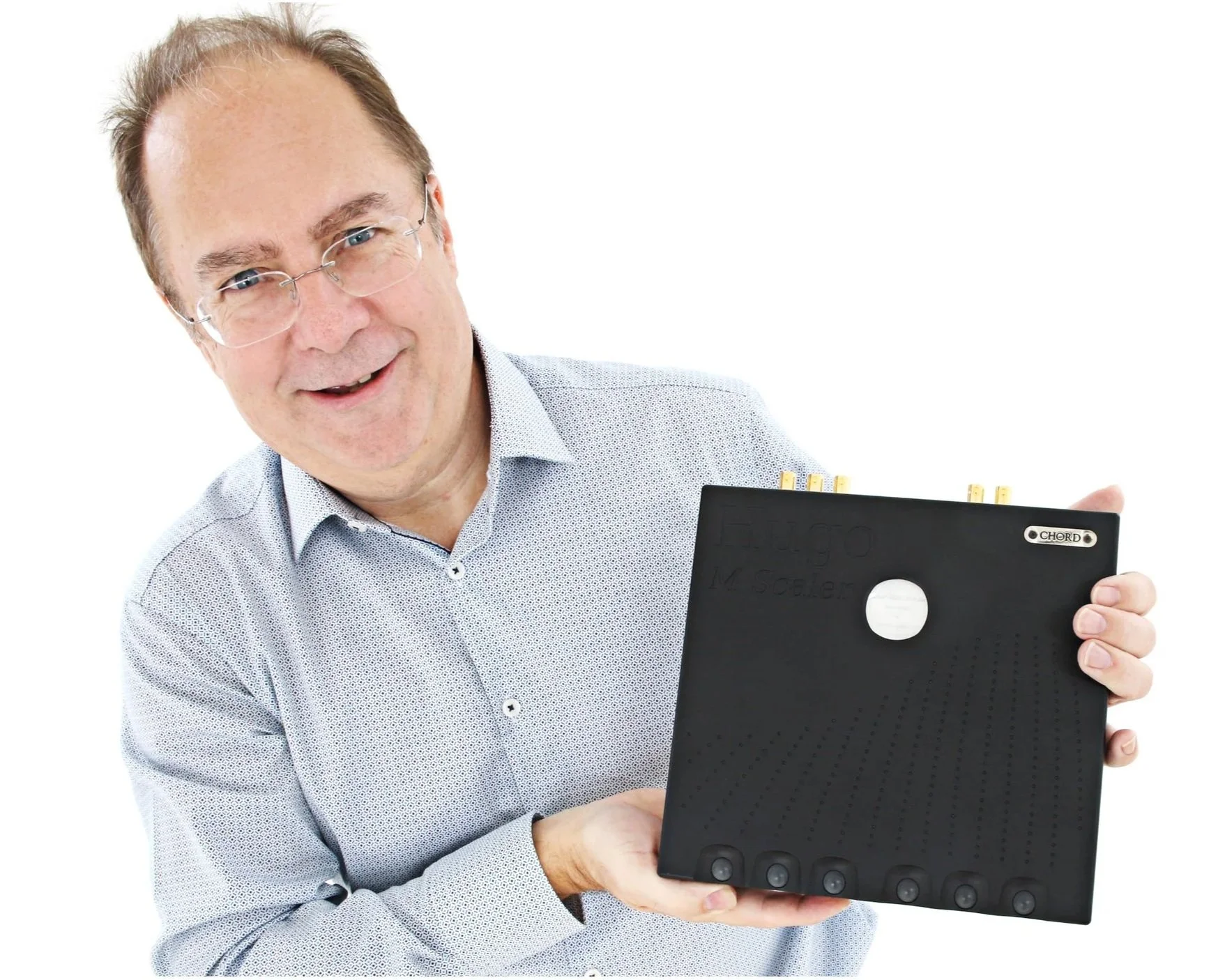 A middle-aged man with glasses, smiling, holds a black electronic device resembling a tape recorder or a music keyboard. The device has a white circular logo and gold-colored connectors on top.