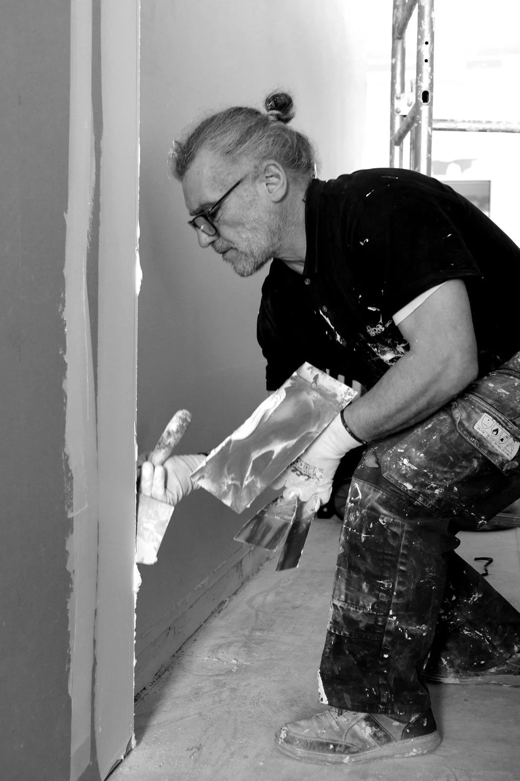Un homme peignant un mur avec un pinceau, portant des vêtements de travail tachés de peinture, en position accroupie.