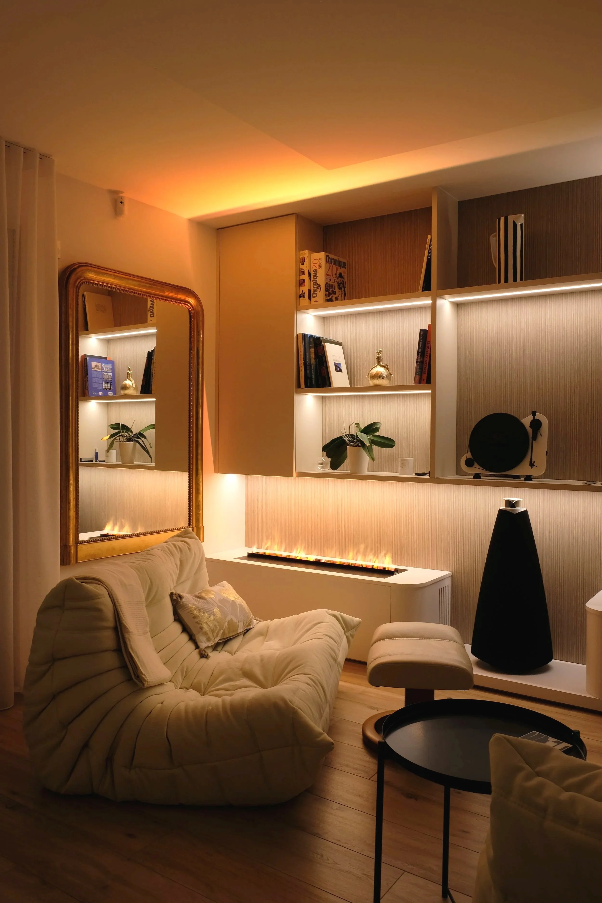 Un salon moderne et cozy avec un canapé beige, un astro lounge noir, une étagère avec des livres et des décorations, un miroir ancien, une plante verte, un mobilier blanc, et un éclairage chaleureux