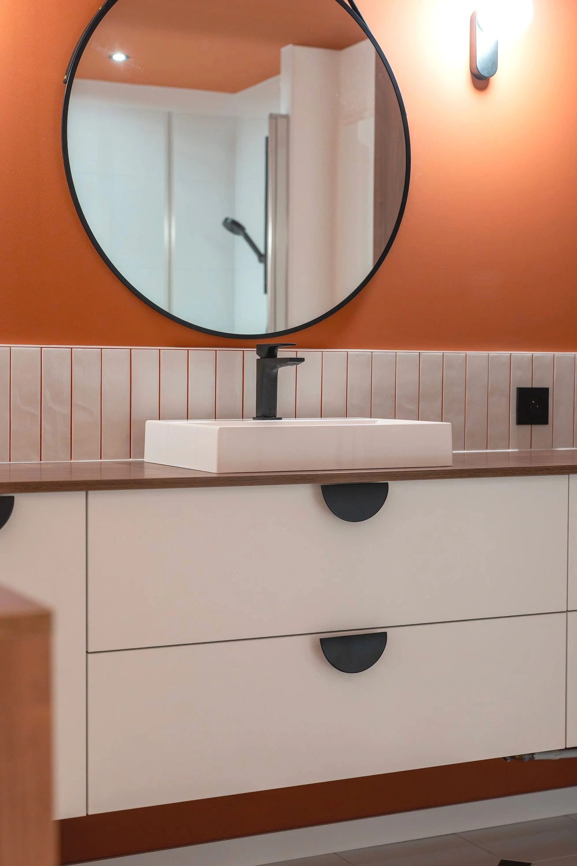 Miroir rond au-dessus d'une vasque blanche avec un robinet noir, le mur derrière est orange avec une section de carreaux blanc et une prise électrique noire, dans une salle de bain moderne.