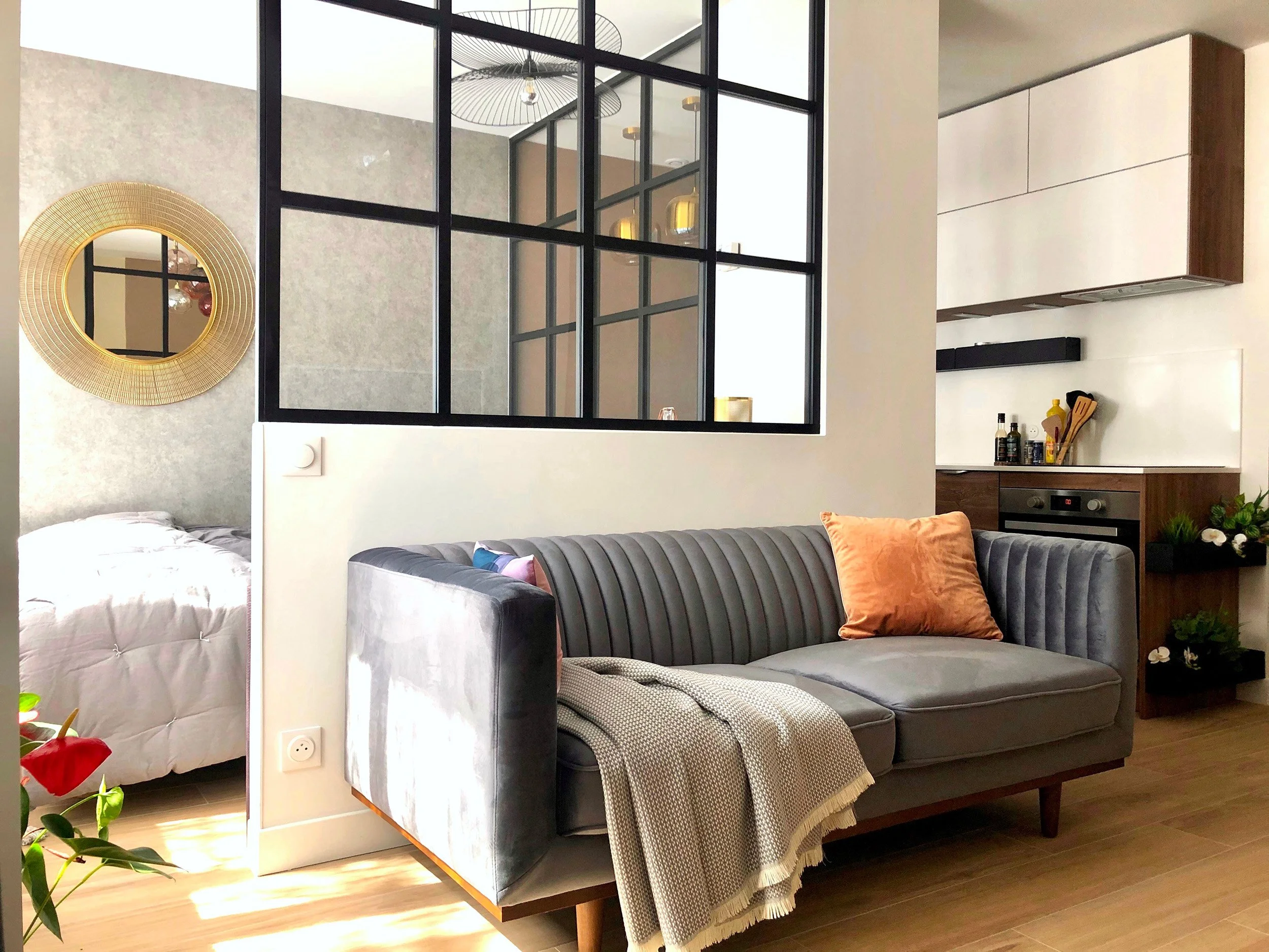 Salon moderne avec un canapé gris, un rideau dans le même ton, des coussins orange, et une cuisine en arrière-plan avec des étagères, des bouteilles et des ustensiles. Une décoration murale ronde dorée est visible, et une séparation en verre noir divise l'espace.