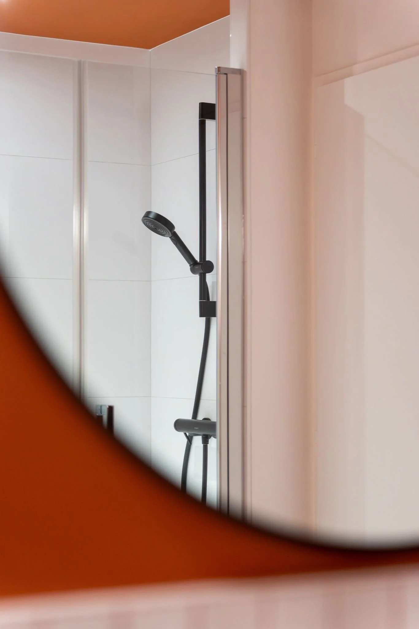Vue d'une cabine de douche avec un pommeau de douche noir et un mur blanc, vue partielle à travers une ouverture encadrée en bois.