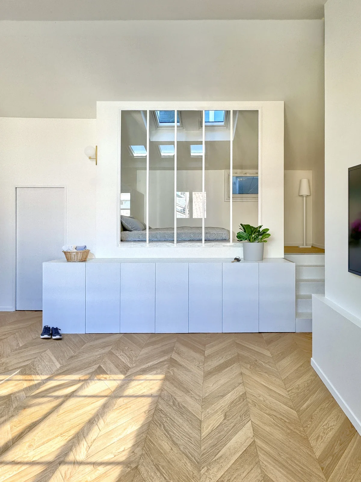 Intérieur moderne avec un mur blanc, un meuble blanc bas, une plante verte, un miroir ou une séparation en miroir, et une chambre avec un lit, vue à travers la séparation. Le sol en parquet en chevrons et quelques chaussures en bas à gauche. Le mur en arrière a des fenêtres de toit.