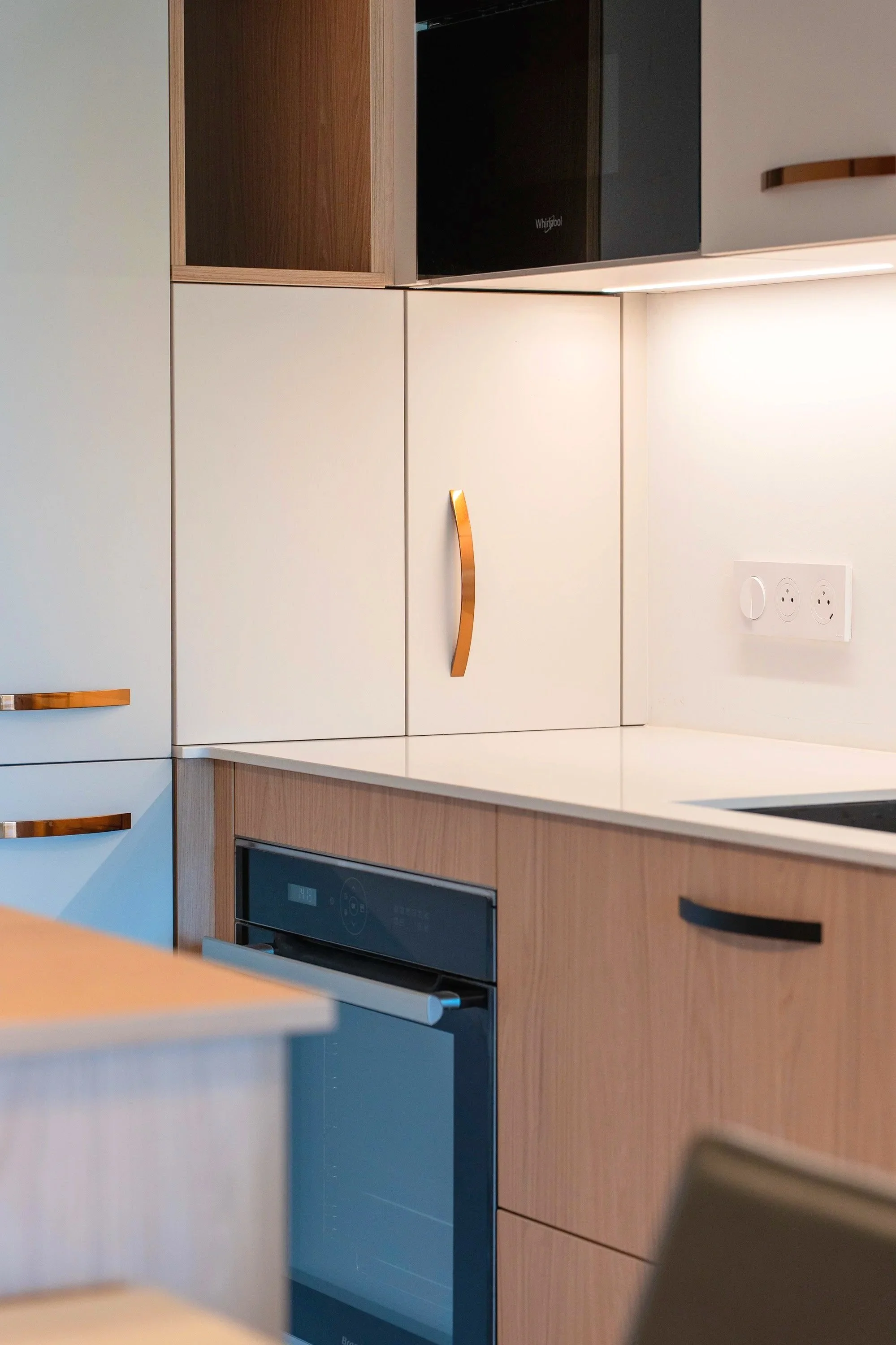 Vue d'une cuisine moderne avec des placards en bois clair, des poignées en cuivre, un four électrique encastré et une prise électrique sur le mur blanc.