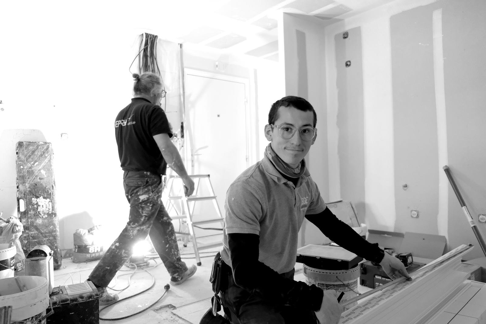 Deux hommes travaillant dans un espace en rénovation, avec des outils et matériaux autour, un homme assis à une table regardant l'objectif, et l'autre debout avec des vêtements de peinture.