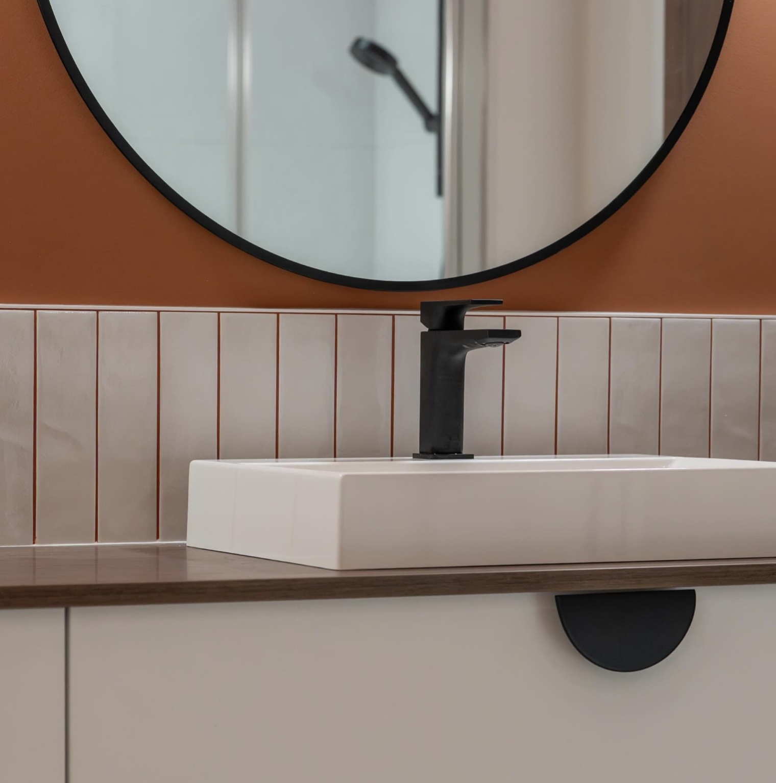 Un lavabo blanc avec un robinet noir, un miroir rond et des carreaux beige et blancs au mur.