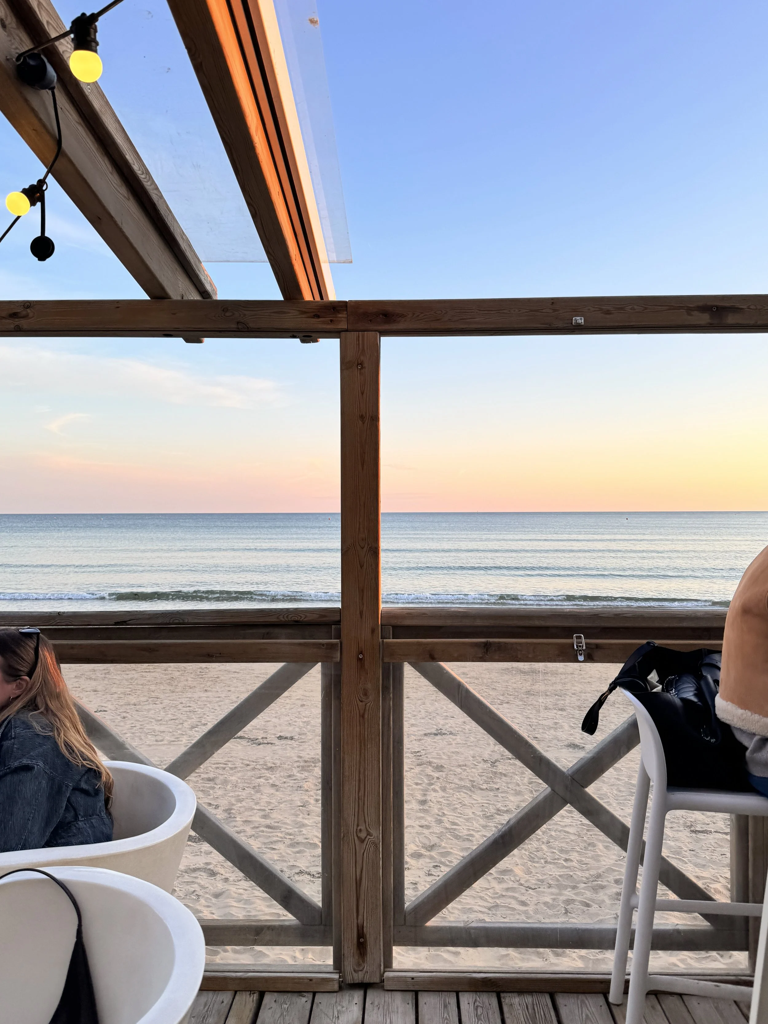Vue de la plage depuis une terrasse en bois, avec des chaises et des tables en plastique blanc, et une structure en bois avec des lumières décoratives, au coucher du soleil.