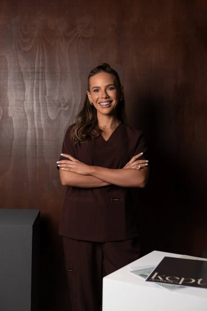 Beth Harrison from Kept Aesthetics wearing chocolate brown scrubs, smiling with arms crossed, standing in front of a dark wooden wall.