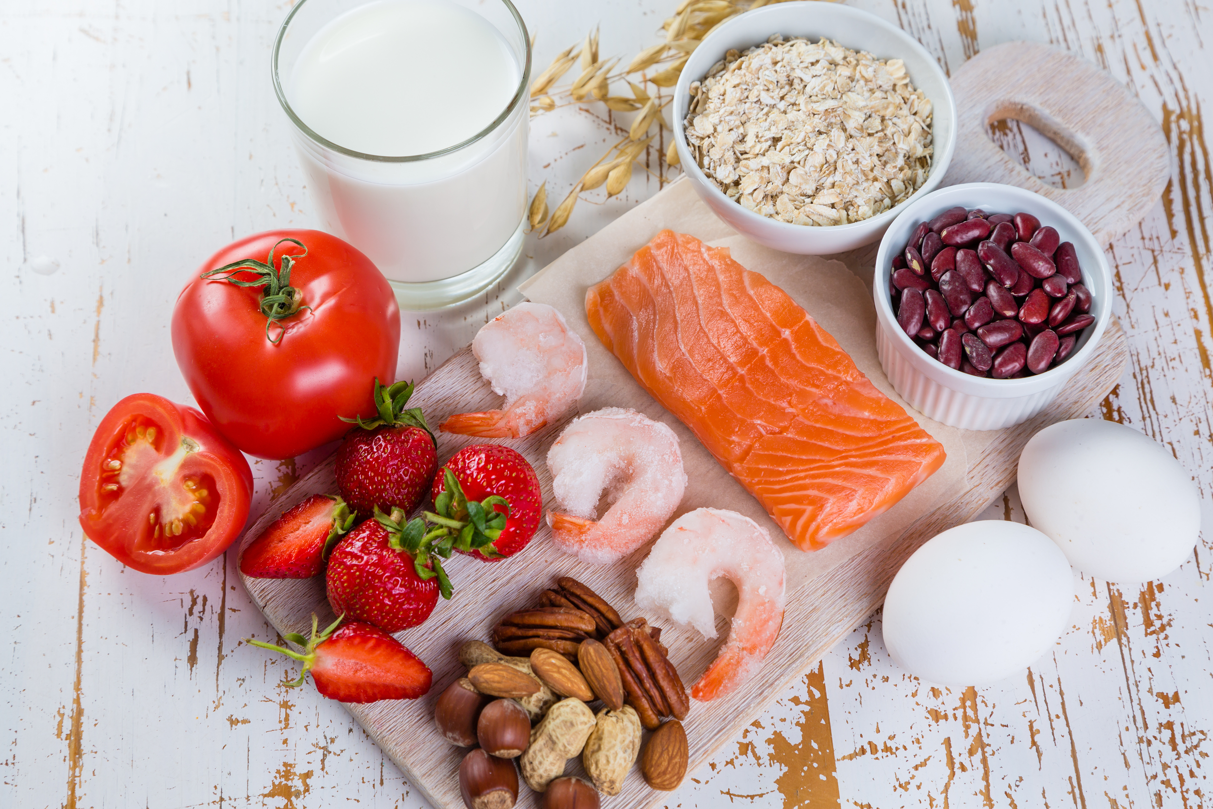 Potential allergens including fresh strawberries, sliced tomatoes, a piece of raw salmon, a bowl of oats, a bowl of red beans, a glass of milk, frozen shrimp, a few eggs, some nuts, and some pecans.