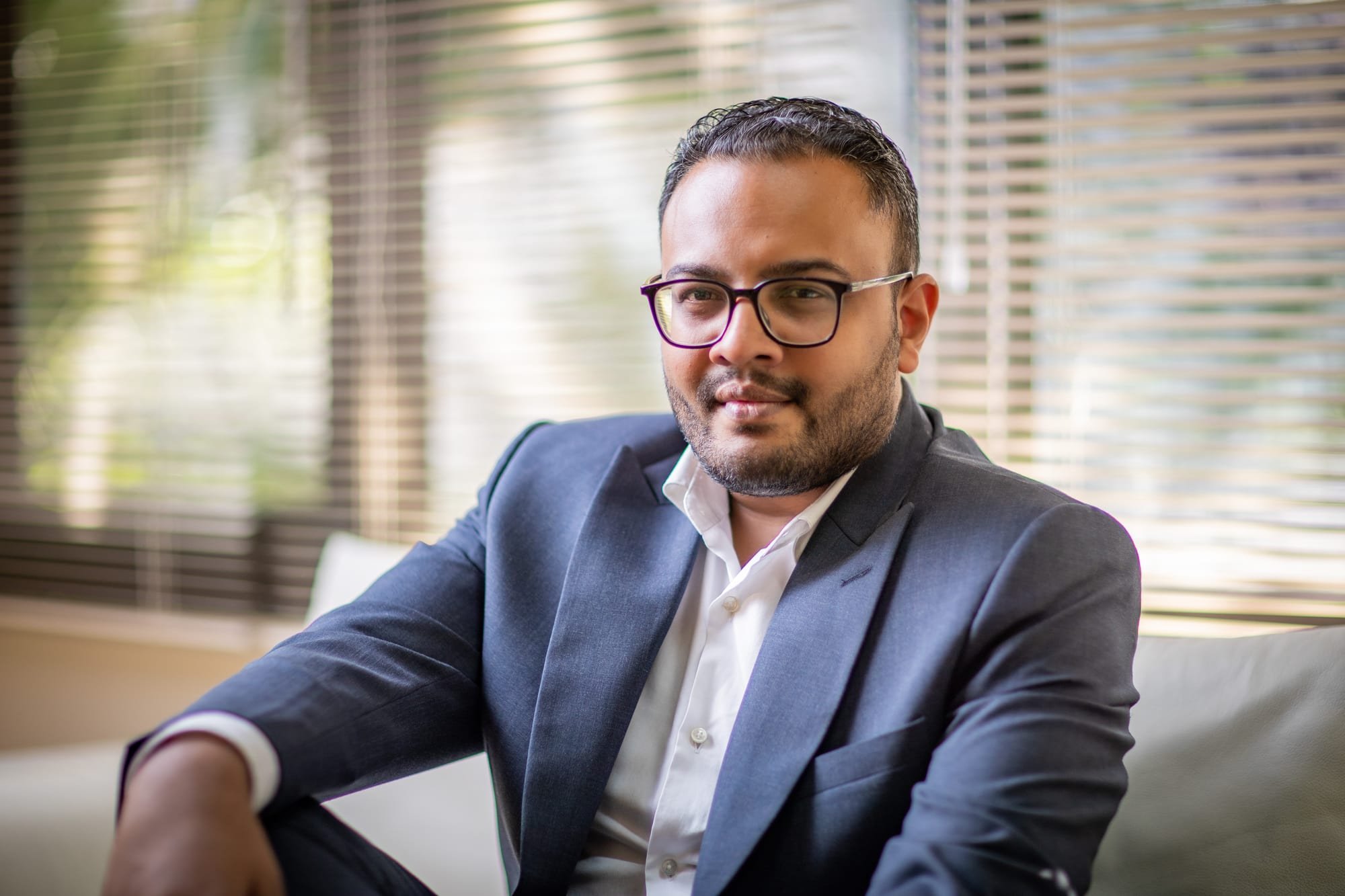 Business portrait of a man wearing glasses and a navy blazer seated beside office window blinds.