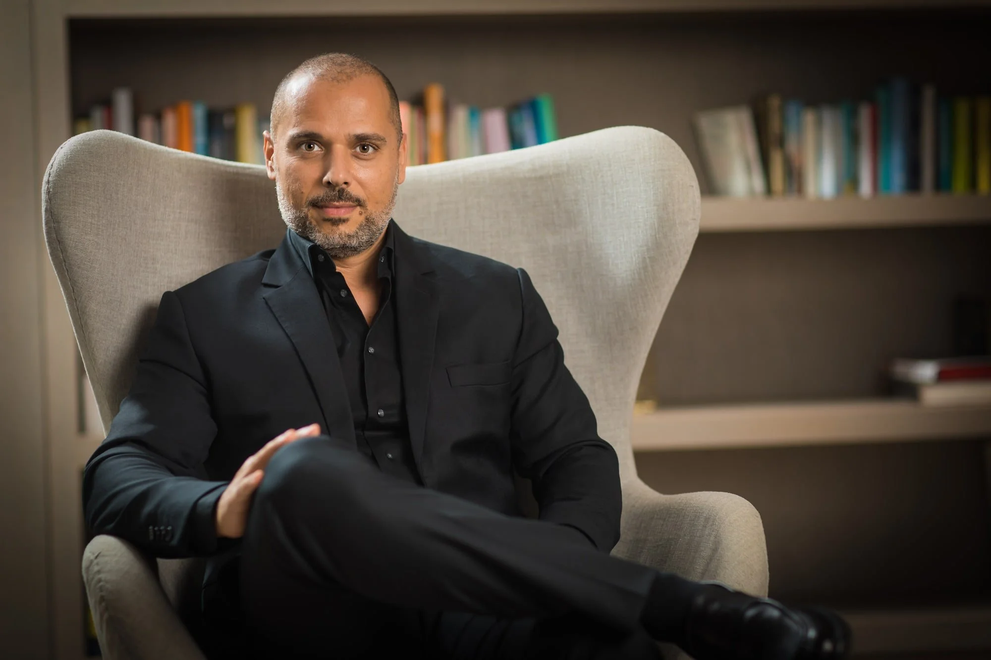 Corporate portrait of Samer Kassem in a dark jacket seated in an armchair with bookshelves behind him.