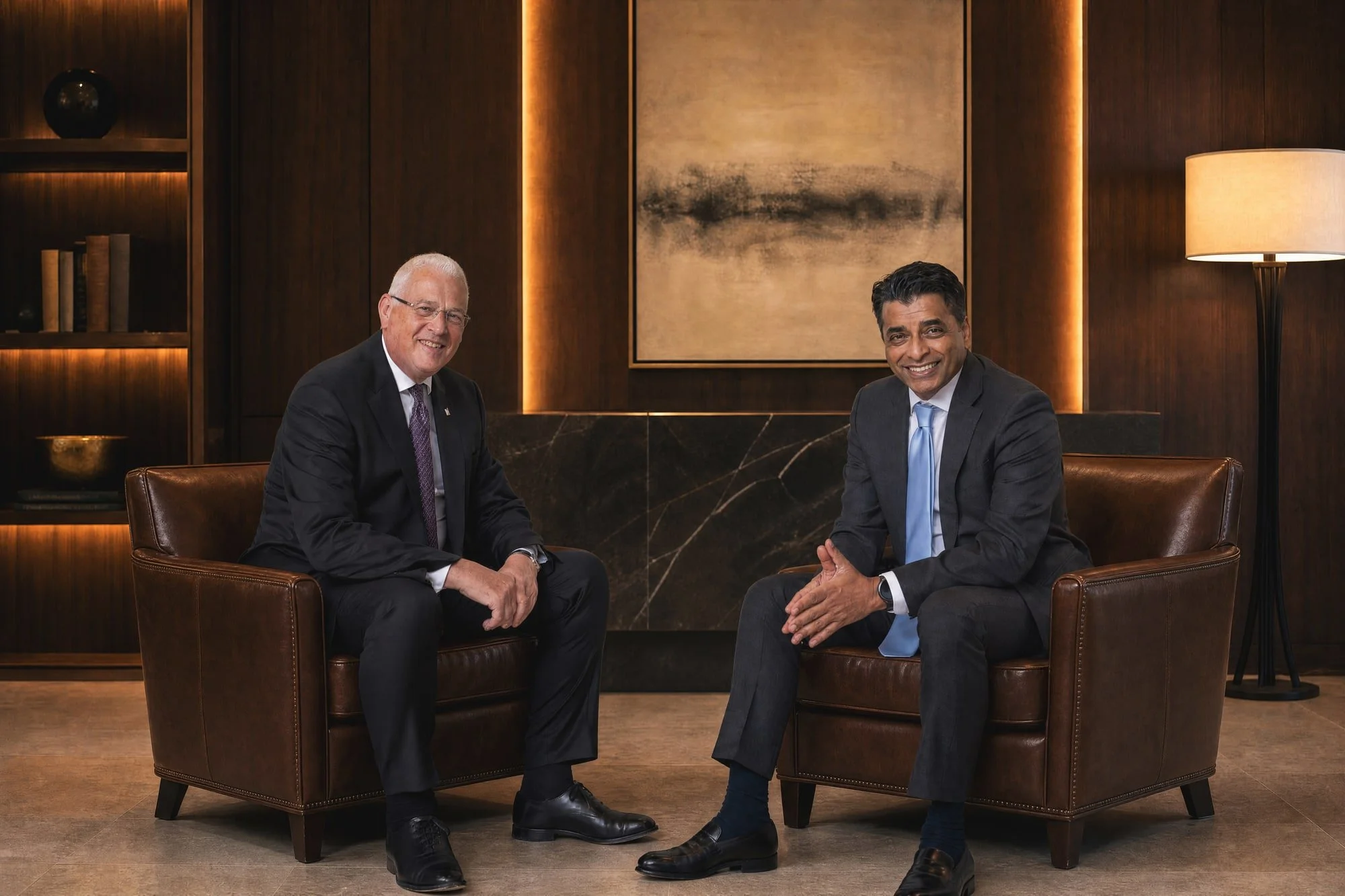 Corporate group portrait of two men in suits seated together in a lounge with framed artwork.
