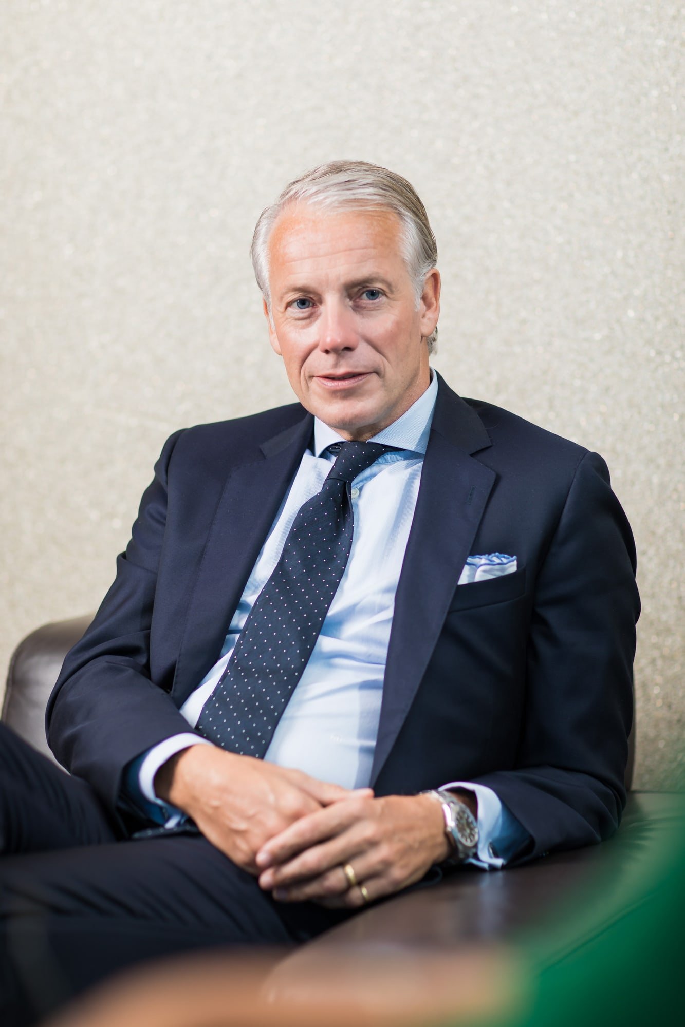 Corporate portrait of a man in a dark suit and tie seated in an armchair against a light background.