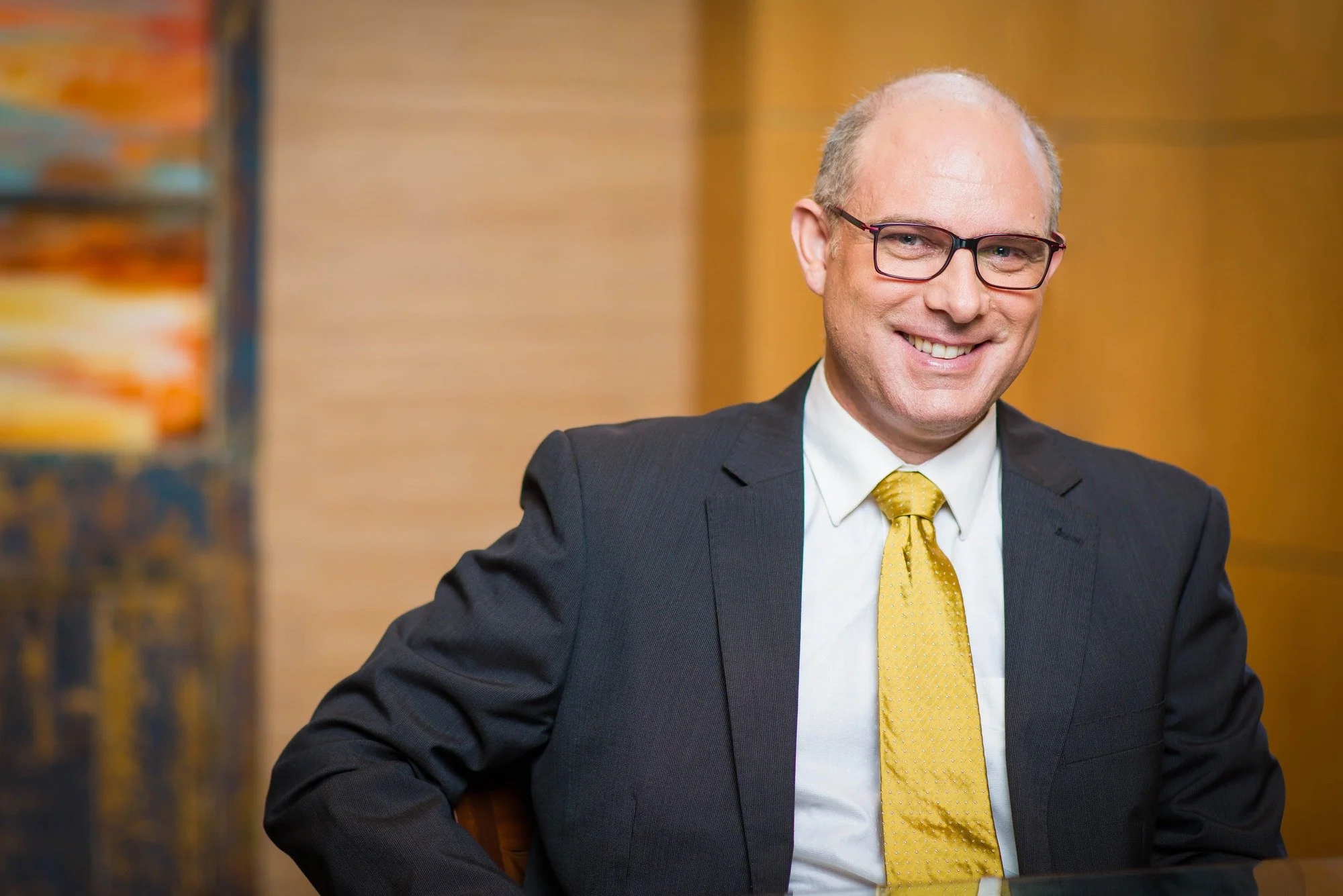 Corporate portrait of Vincent Noël wearing glasses, a dark suit and a yellow tie, seated against wood panelling.