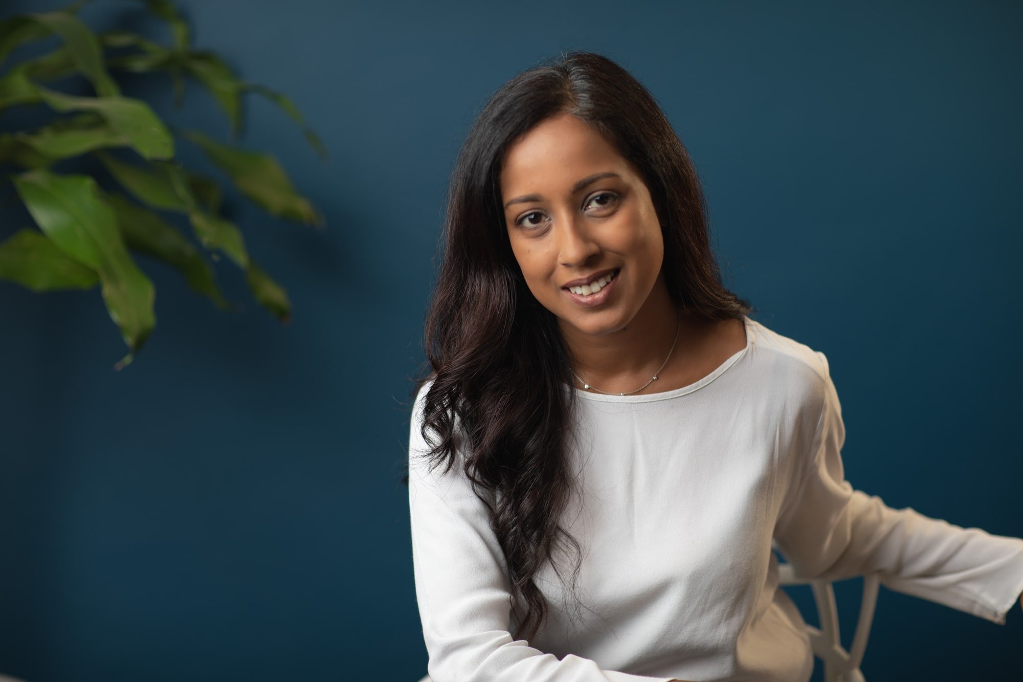 Business portrait of a woman in a light blouse seated against a blue office wall.