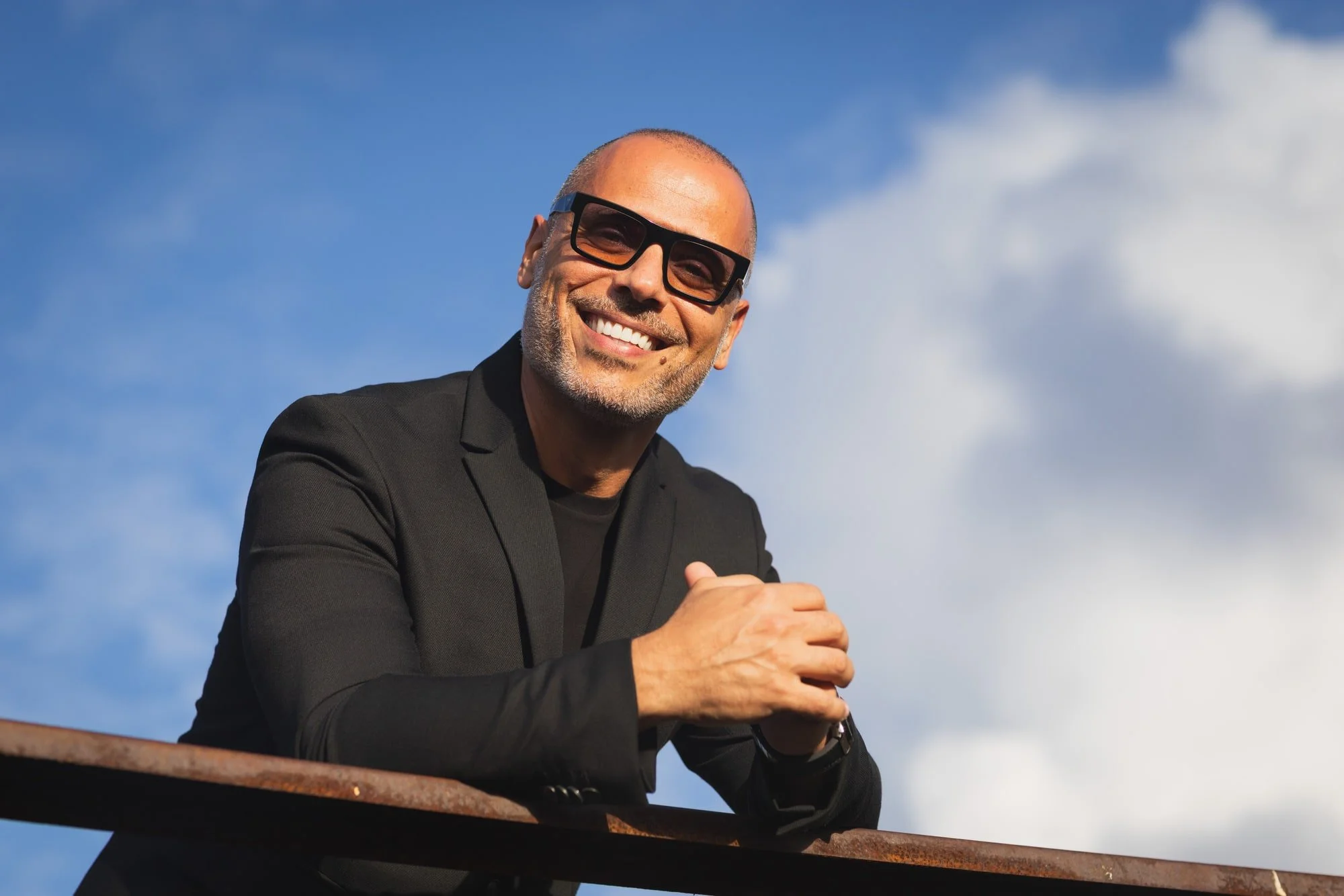 Outdoor portrait of Samer Kassem in a black shirt and sunglasses against a blue sky.