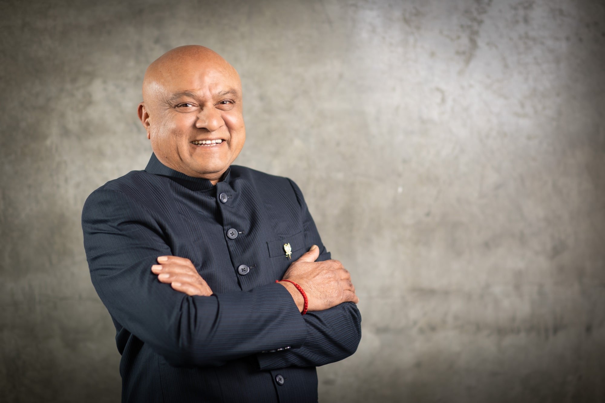 Professional portrait of a smiling man in a dark shirt with folded arms against a textured grey background.