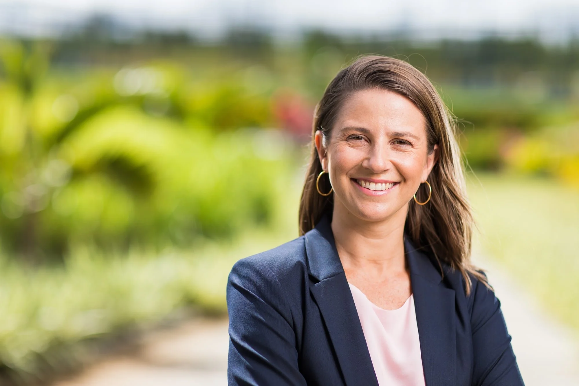 Outdoor professional headshot of Jessica Valet in a navy blazer with a softly blurred green background.