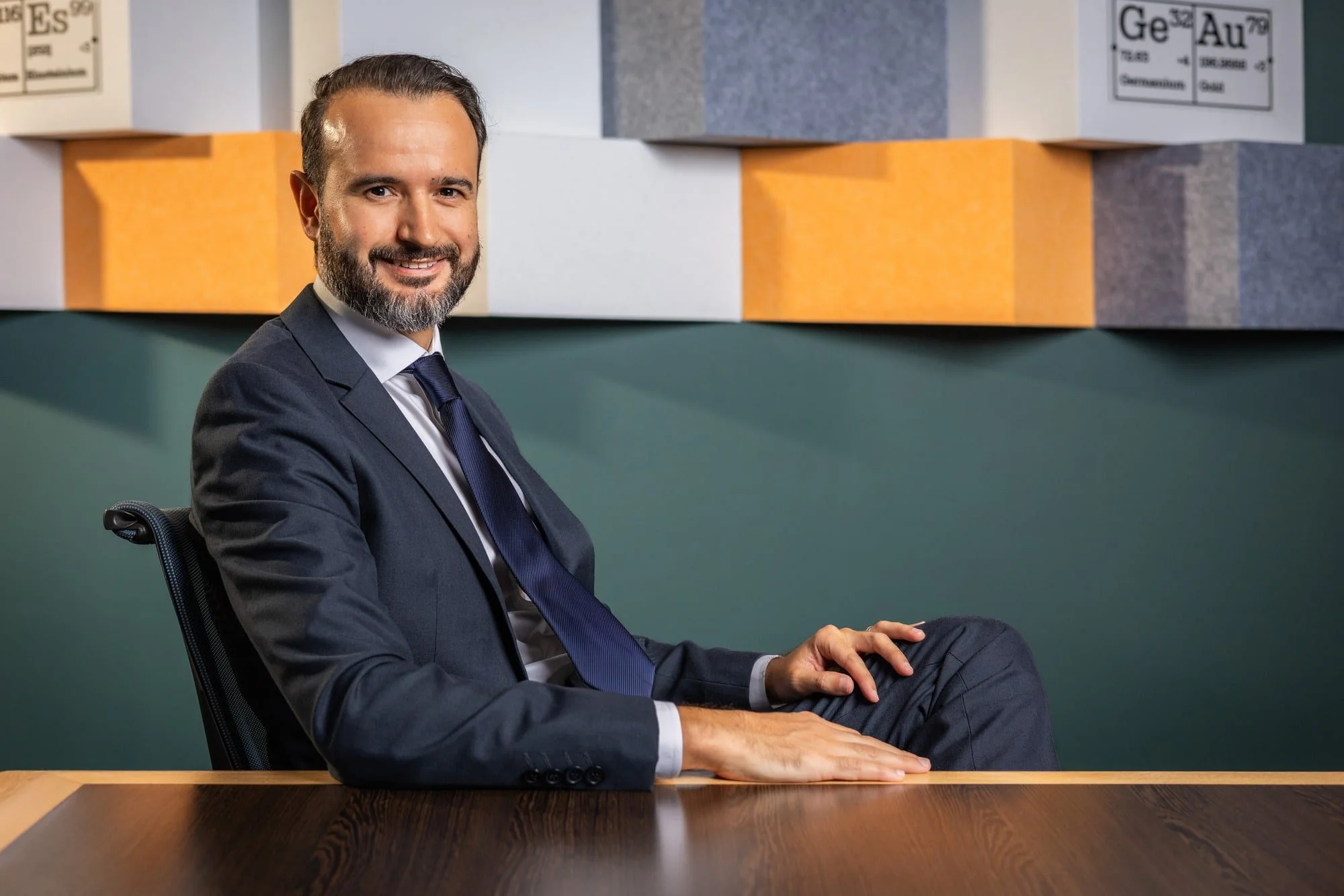 Professional portrait of Jonathan Wienand in a dark suit seated at a table beneath geometric wall artwork.