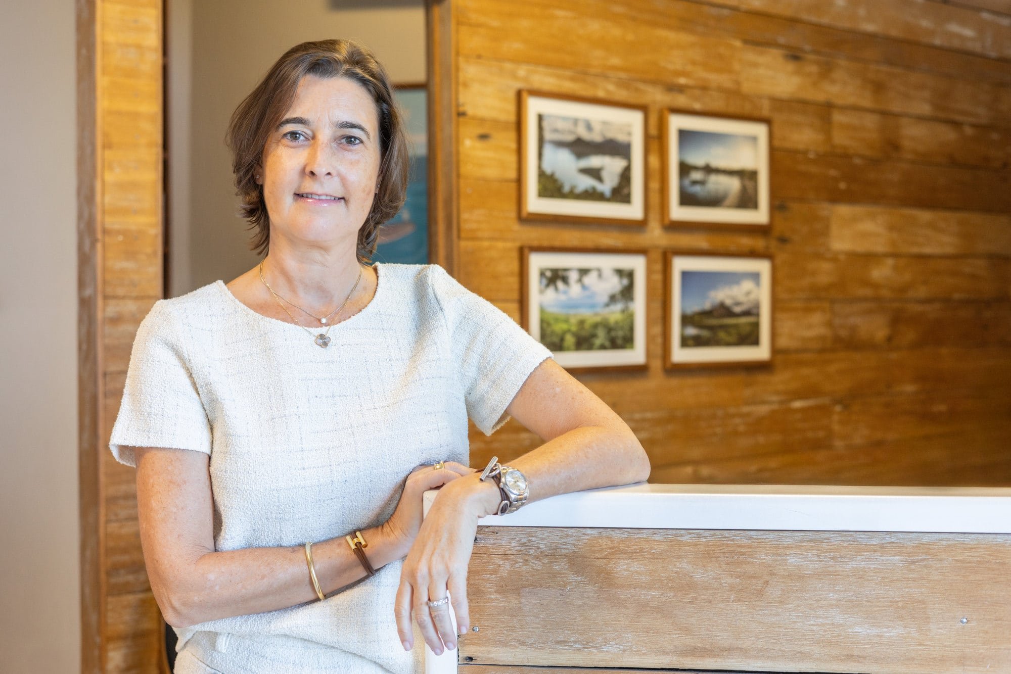 Professional portrait of Frédérique De Beer in a white top leaning on a counter beneath framed photographs.