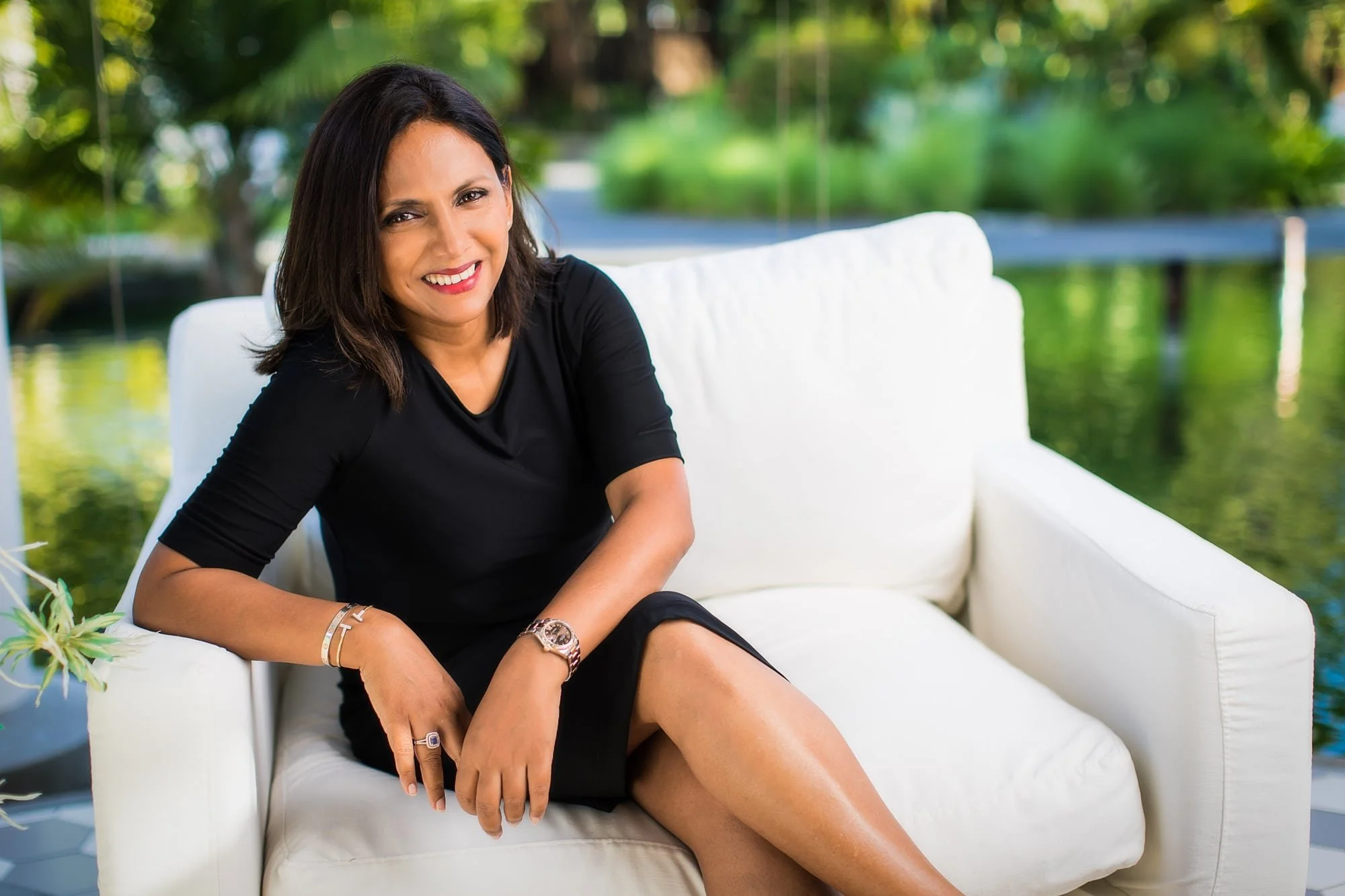 Professional portrait of Aisha Aly in a black dress seated on a white sofa overlooking a garden.