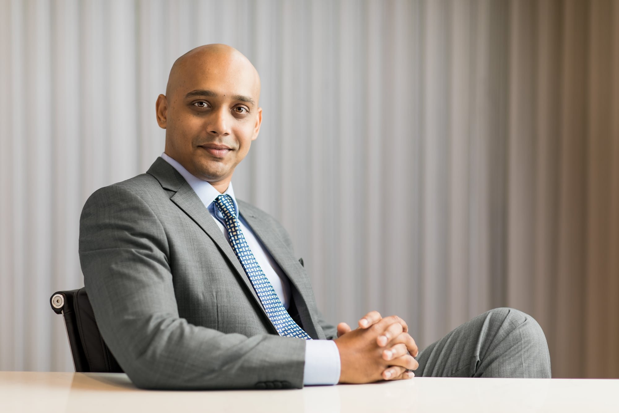 Corporate portrait of a man in a grey suit seated beside softly lit curtains.