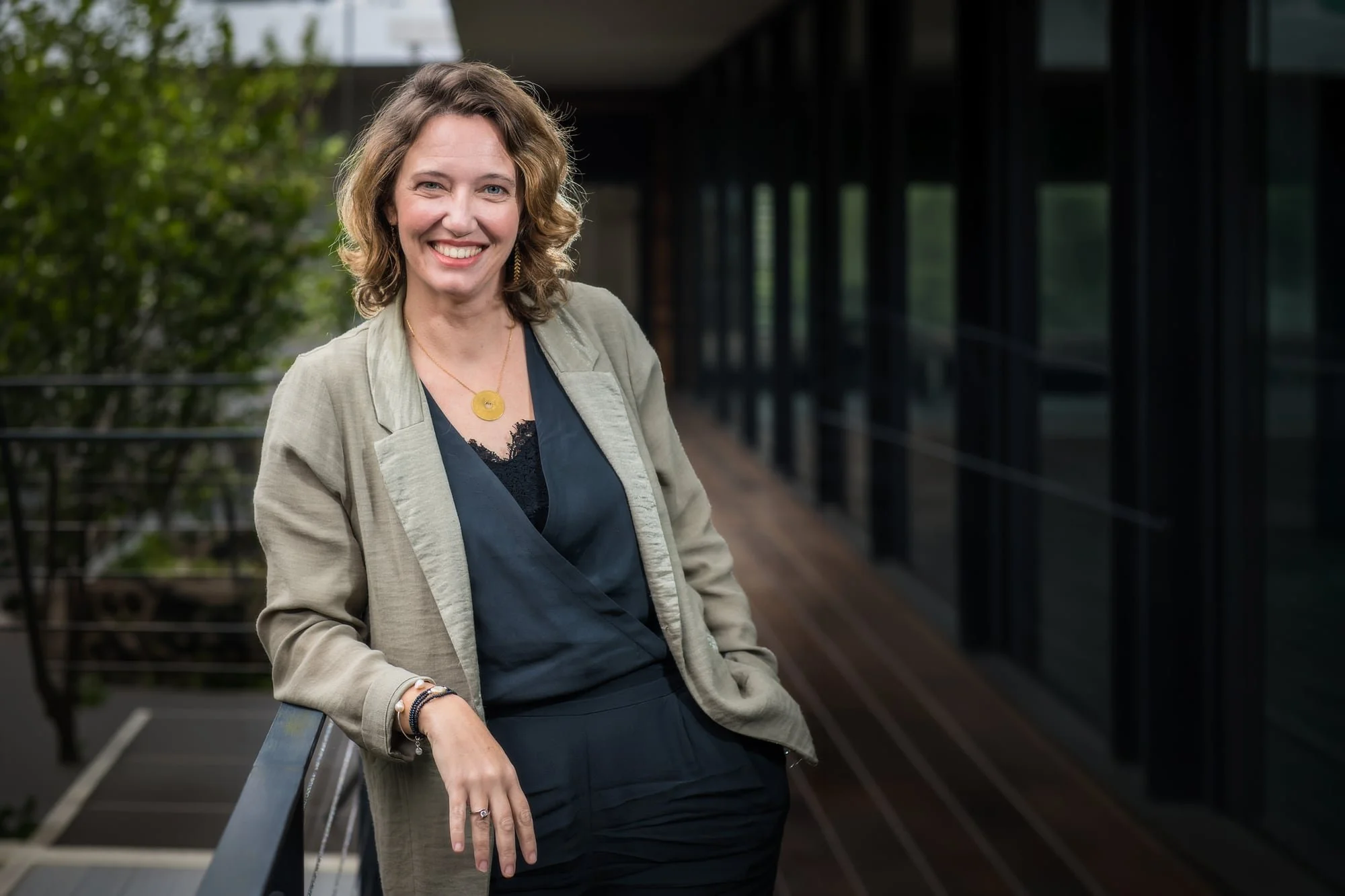 Outdoor business portrait of a woman in a beige blazer beside a modern building and greenery.