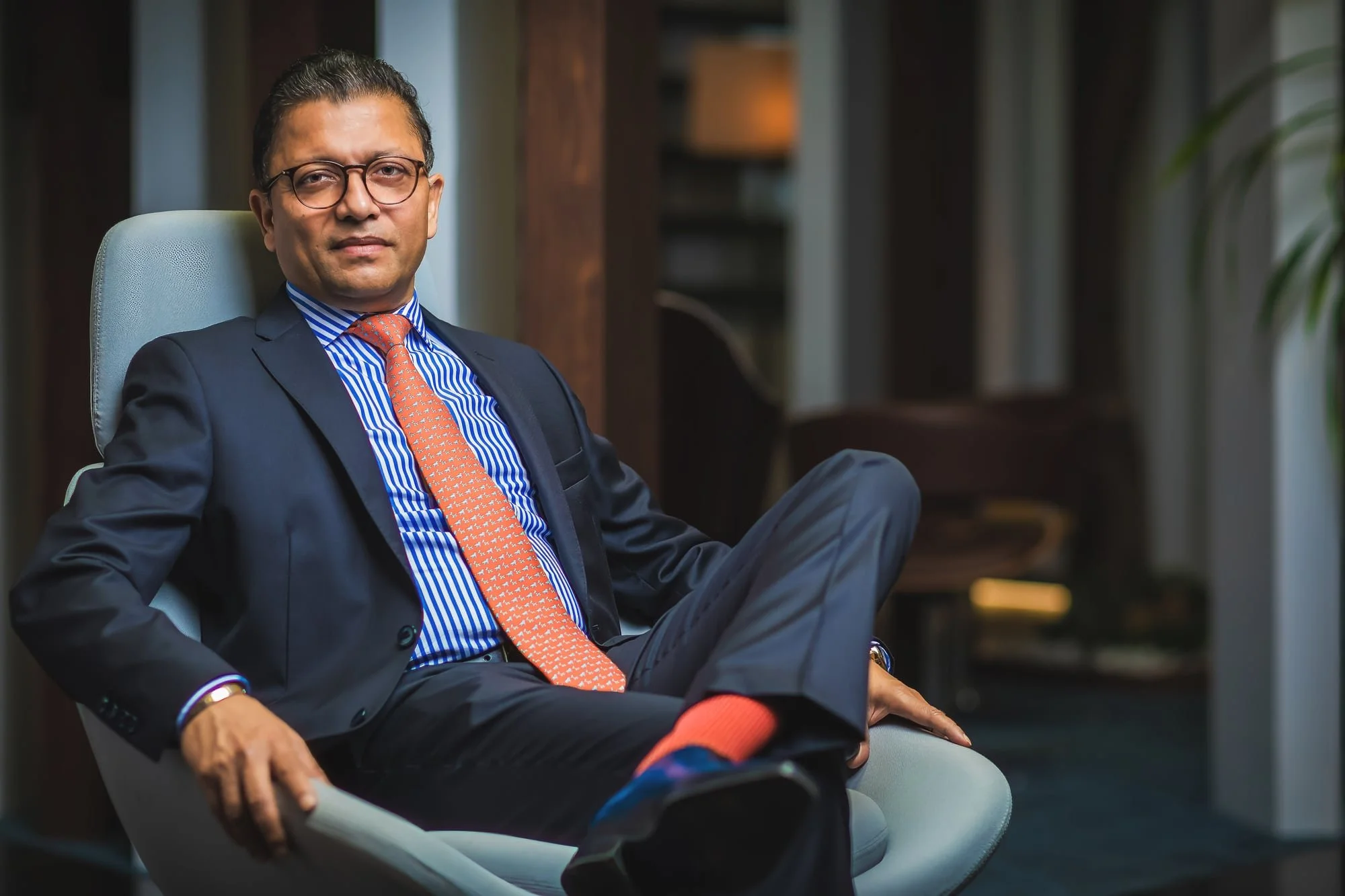 Corporate portrait of a man in a navy suit and orange tie, seated in a light blue armchair in a contemporary office.