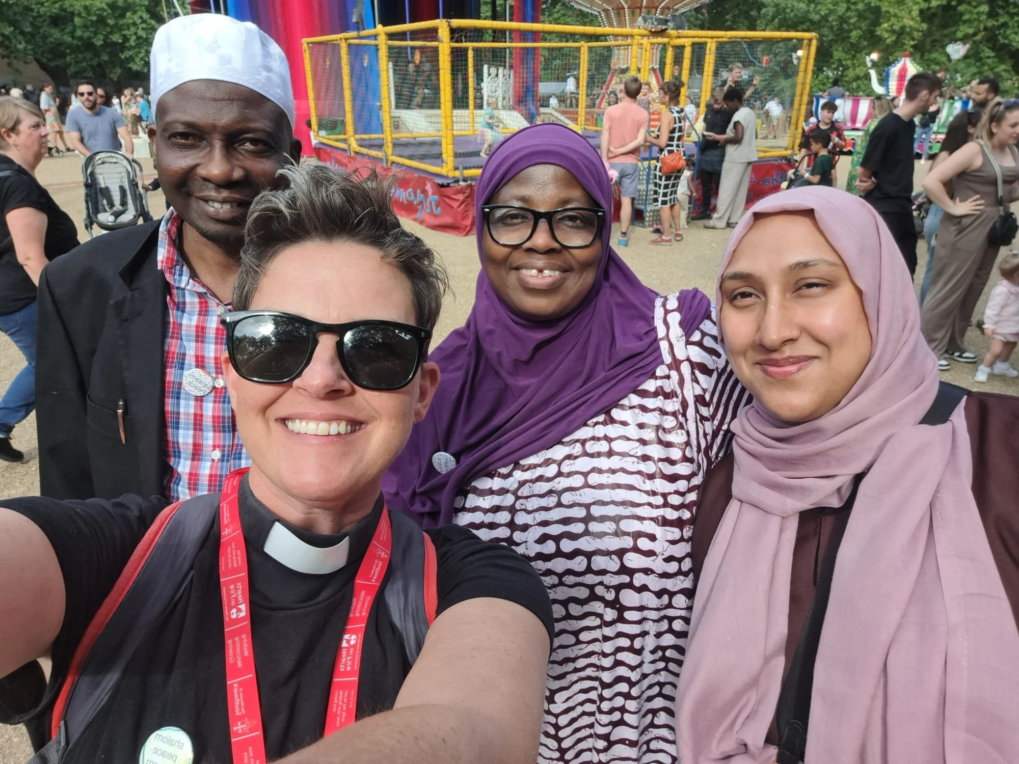 A group of four diverse people, two women and two men, taking a selfie at an amusement park, with a colorful ride and crowds in the background.