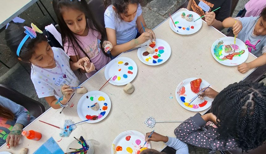 Children painting on rocks with paint and brushes around a table.