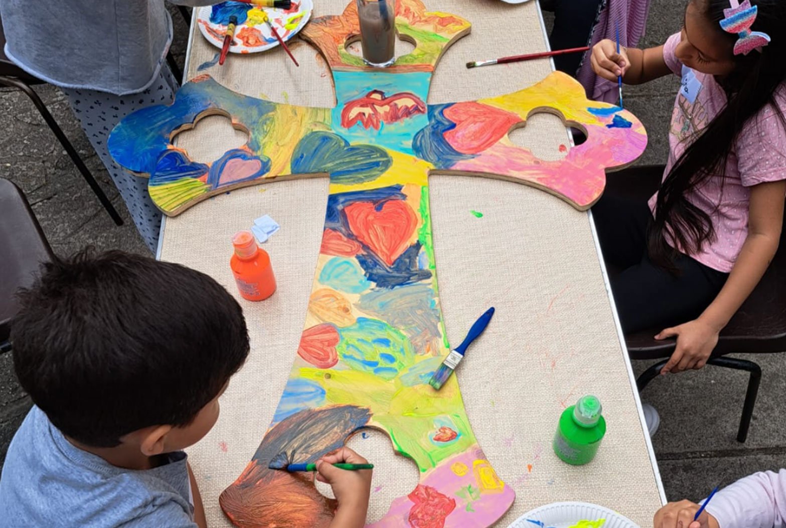 Children painting a large cross-shaped wooden cutout at a table outdoors, with colorful abstract and heart designs.