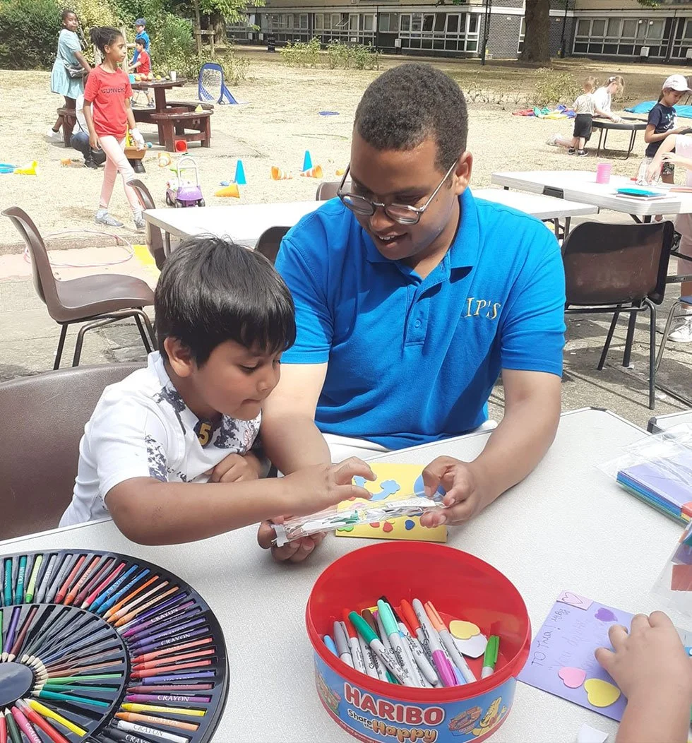 A young boy and a slightly older man, possibly a mentor or teacher, are sitting at a white table outside, engaged in a craft activity with colorful paper and markers. The boy is reaching for a craft item while the man is smiling and assisting him. Ot