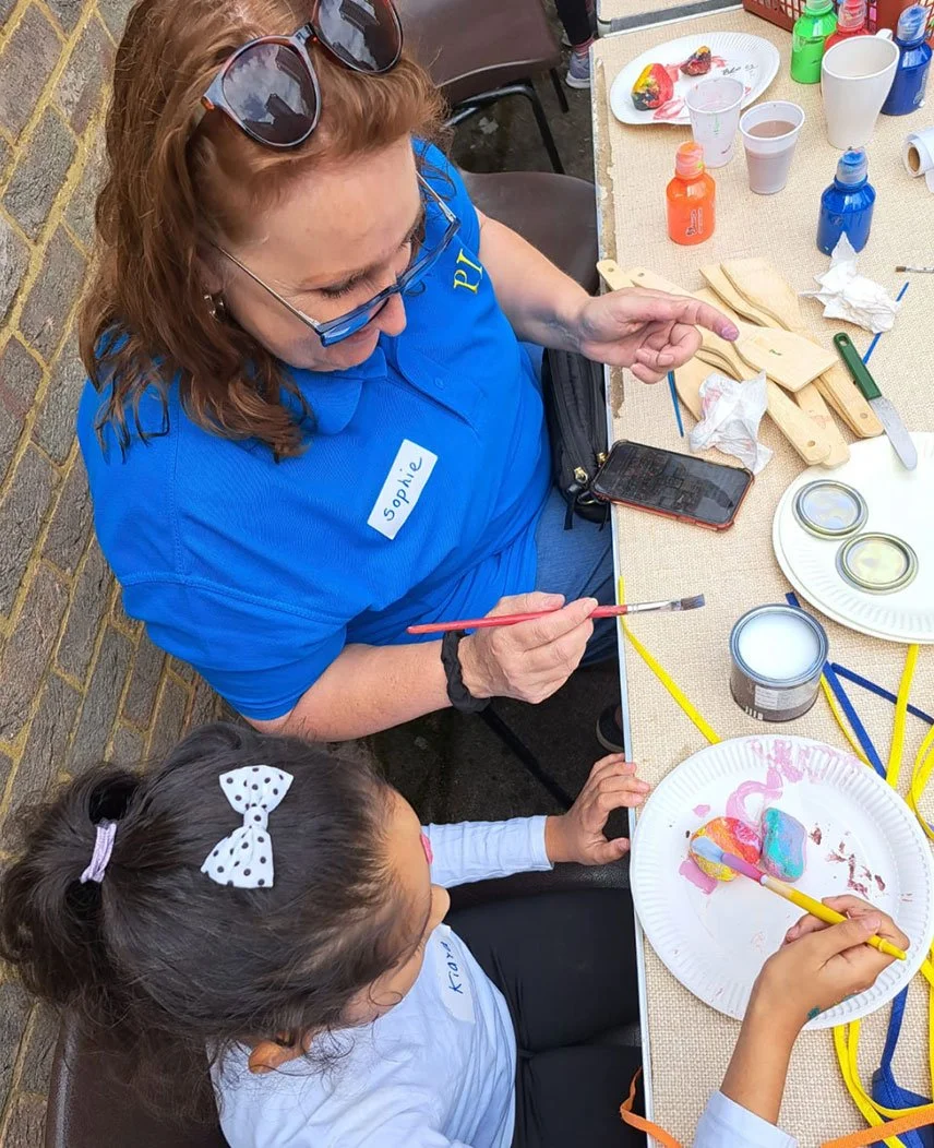 An adult woman and a young girl are sitting at a table, painting and decorating objects with paint. The woman is wearing glasses, a blue shirt with a name tag that says "Sophie," and sunglasses on her head. The girl has a white shirt with a polka dot