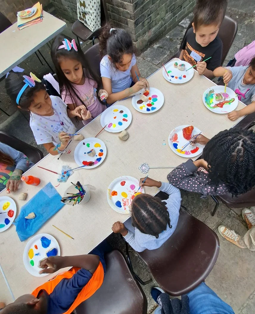 Children sitting around a table painting rocks with colorful paints during an outdoor art activity.
