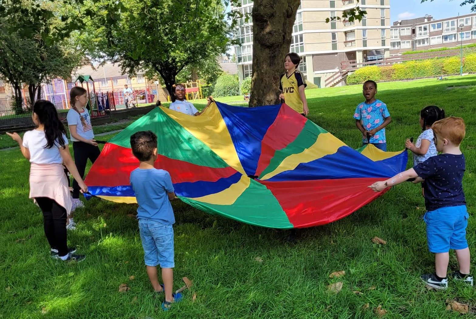 Children playing with a colorful parachute in a park, with trees and buildings in the background.
