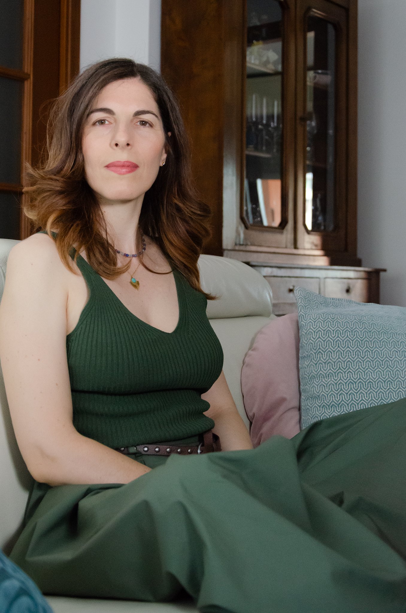 A woman with shoulder-length brown hair wearing a dark green sleeveless top, sitting on a cream-colored couch with decorative pillows, in a room with wooden furniture and a china cabinet in the background.