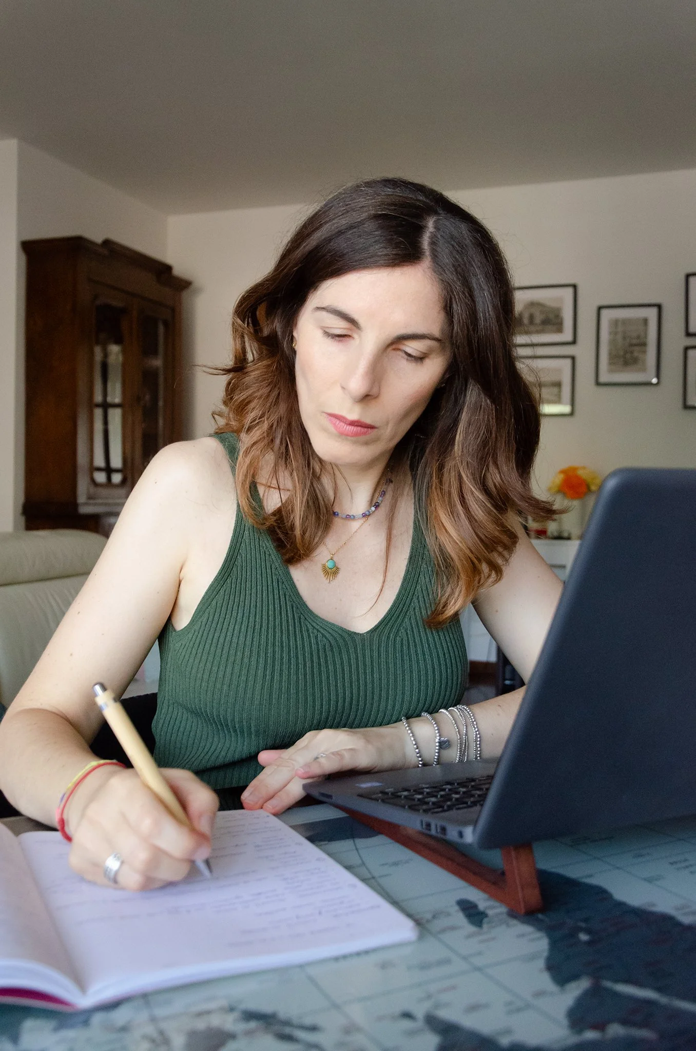 A woman with shoulder-length brown hair wearing a green sleeveless top is sitting at a table, writing in a notebook with a pen. There is a laptop in front of her and she is looking at it. The background shows framed pictures on the wall, a wooden cabinet, and some flowers on a table.