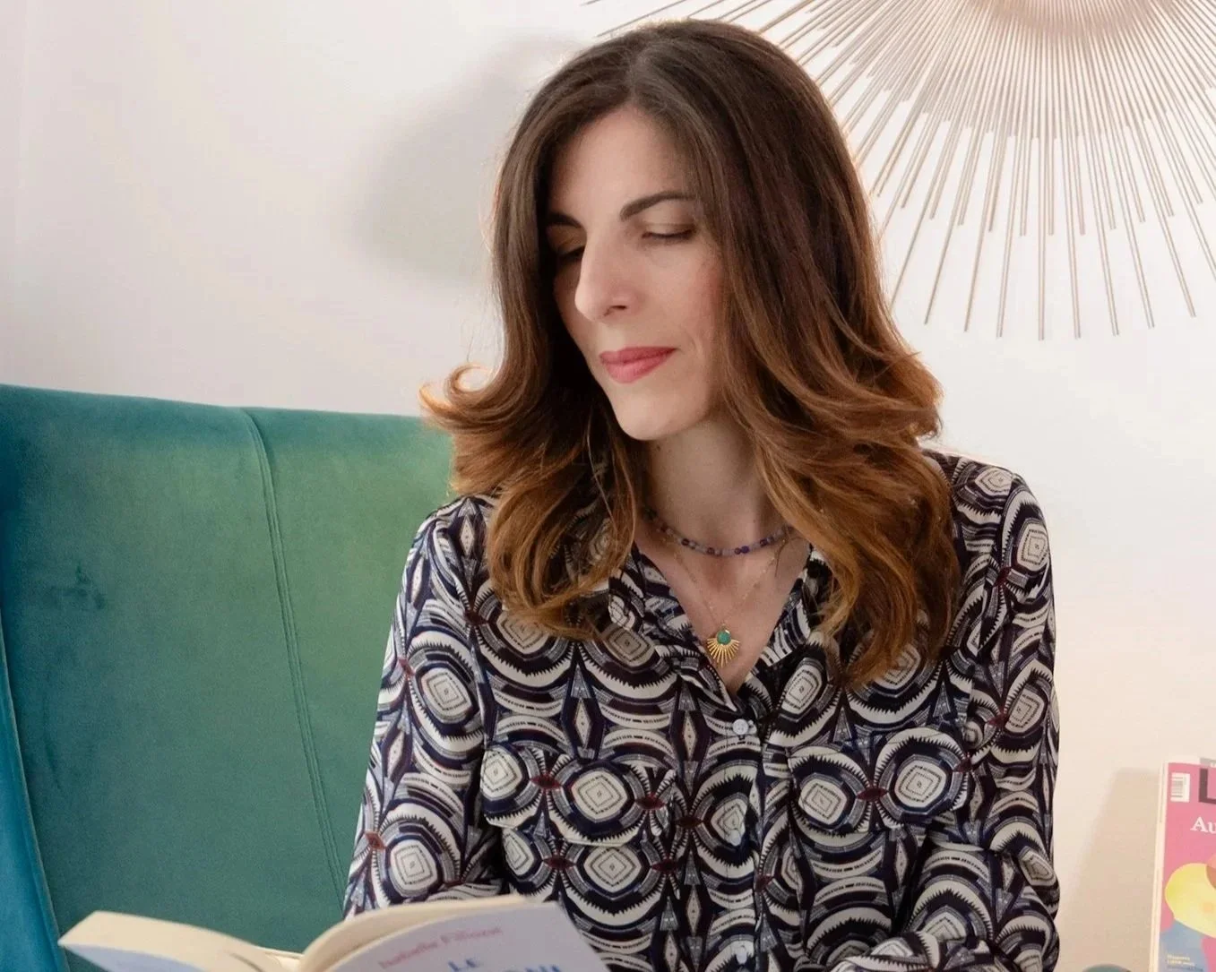 A woman with wavy, shoulder-length brown hair sitting on a green chair, reading a book. She is wearing a patterned blouse and layered necklaces, with a sunburst wall decoration behind her.